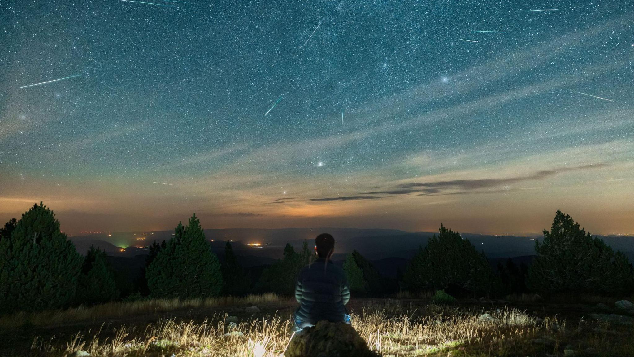 A man sits on top of a mountain watching the Perseid meteor shower
