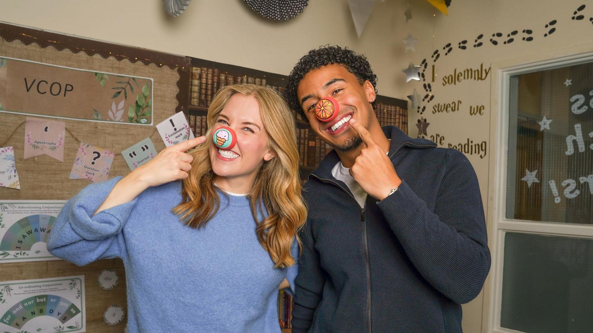 Maddie Moate and Kit Brown smiling at the camera while pointing at their red noses that have pictures on them. Maddie is white, has long blonde curly hair and is wearing a fleecy light blue jumper and blue jeans. Her red nose has an ice cream on it. Kit is black and has short curly black hair and a stubbly beard. He is wearing a navy blue cardigan. His red nose has a flower on it. They are stood in the corner of a classroom with various pinboards, decorations and a window behind them.