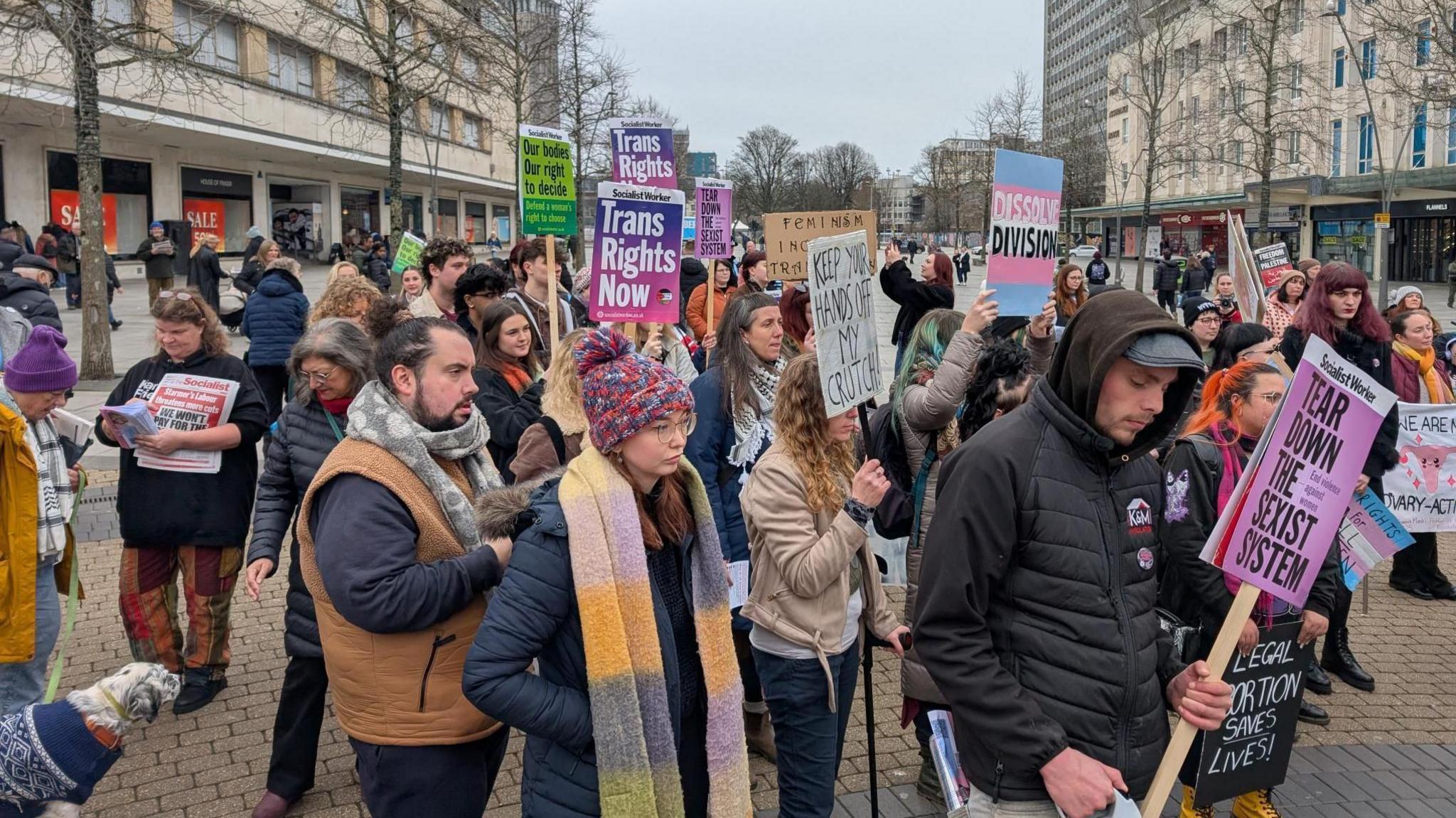 Protestors gather in the city centre. They are dressed in coats, hats and scarves. Some people are holding signs. One sign states 'tear down the sexist system' while another one states 'trans rights now'.