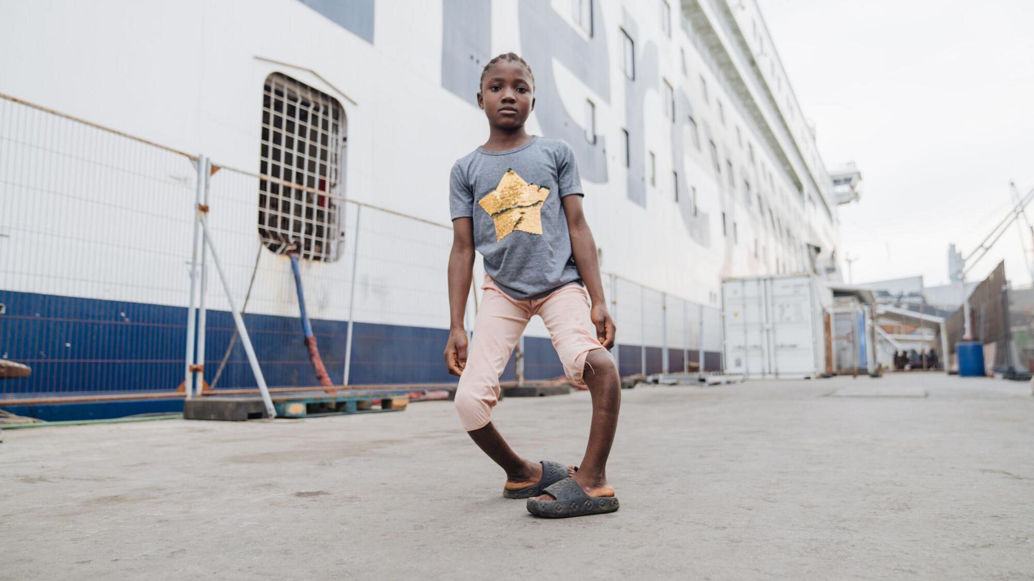 Fudia, before her surgery, she stands outside in a dockyard, with a white and blue Mercy Ship in the background. Fudia wears her hair in braids, with a slate grey short sleeved cotton t shirt and a gold foil star on the front, she has pale pink capri shorts and wears black platform sliders, her legs are severely bowed and the knees, with her feet pointing inwards.