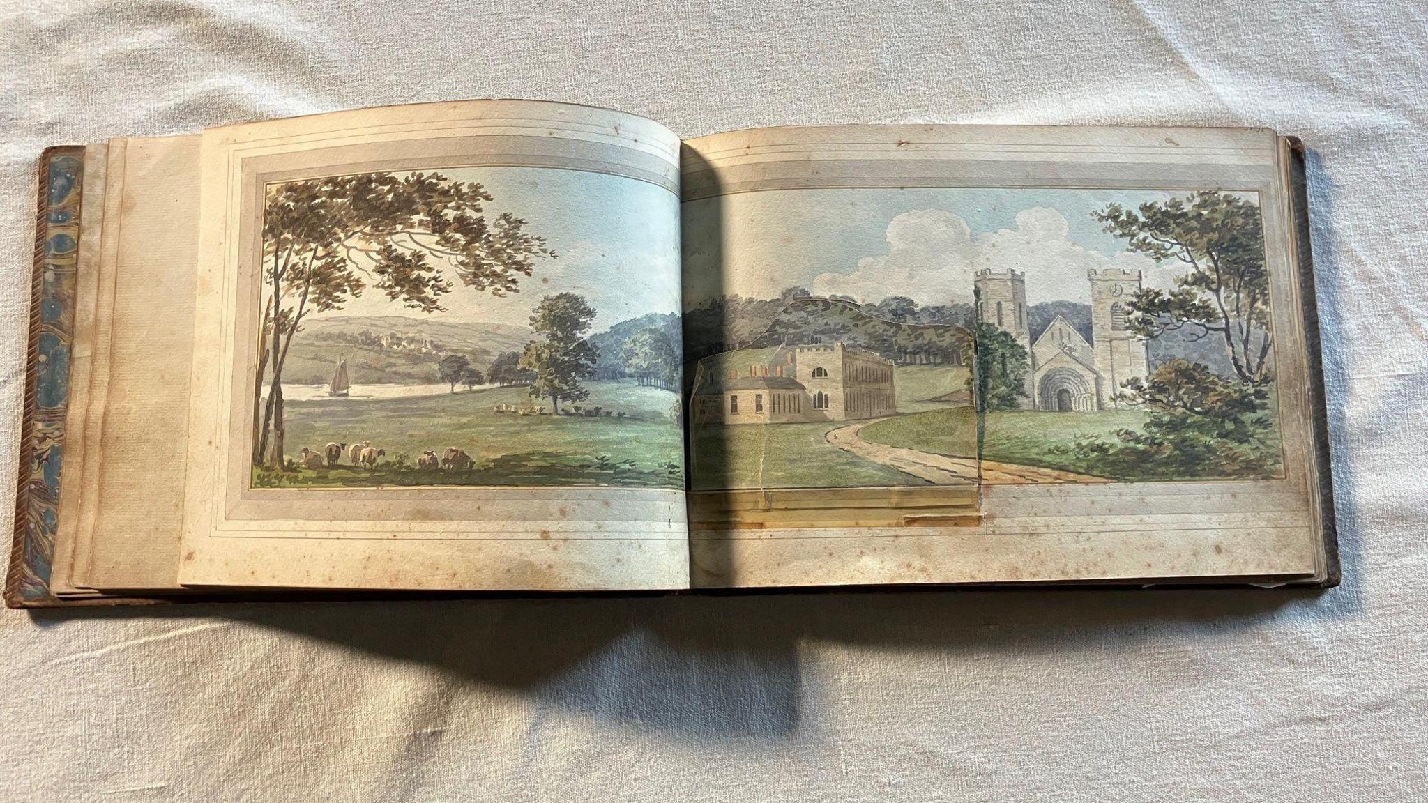 The open Red Book which shows a hand-drawn landscape with a river on the left with a boat on it, trees and then the Port Eliot house and church to the right. The pages are mottled with age.