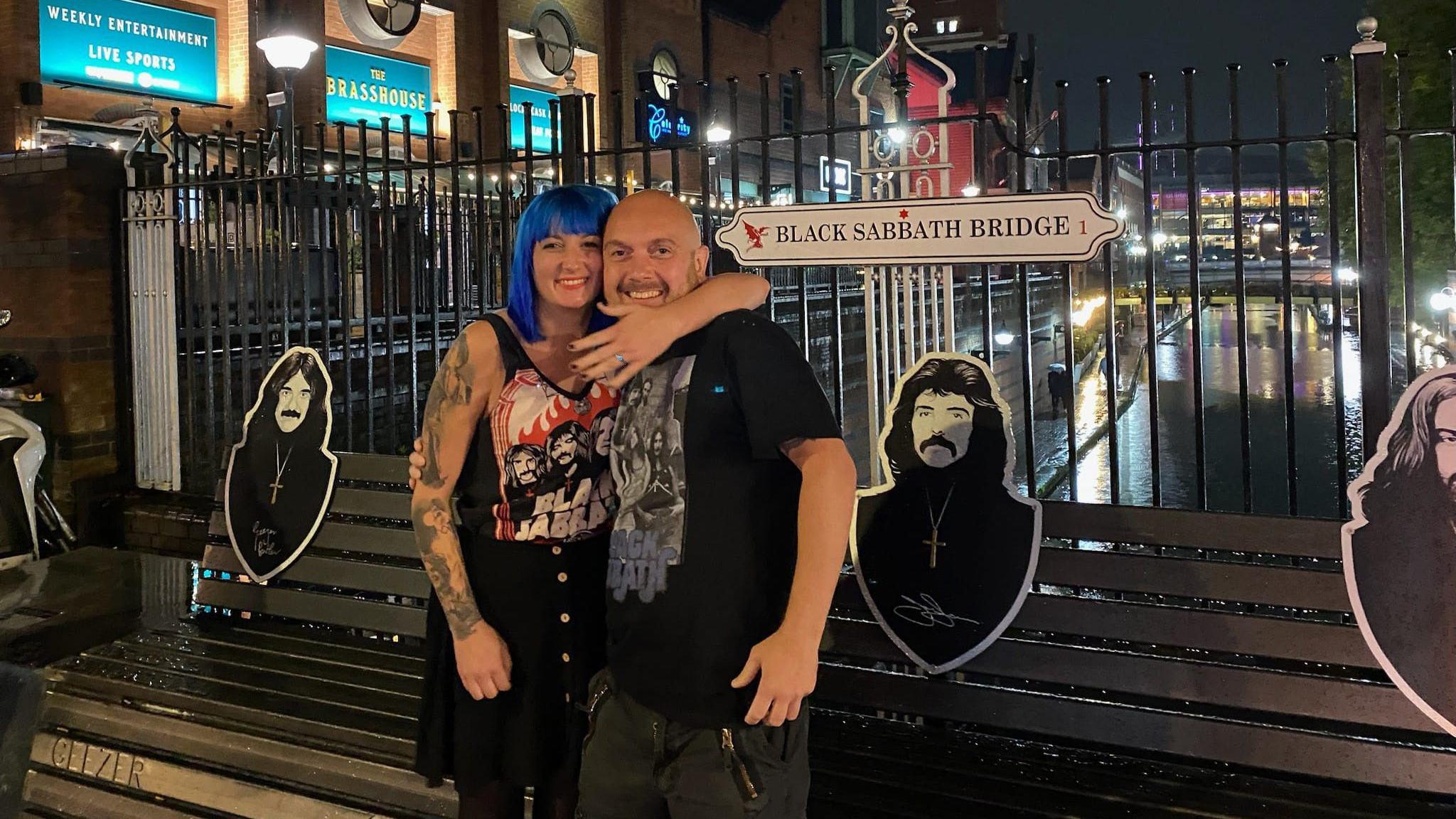 A man and a woman are standing in front of a bench with black and white cut-outs of Black Sabbath members on it. The bridge is in front of a metal fence which overlooks a canal. The woman, with blue hair, is wearing a Black Sabbath t-shirt and has her arm around a man, showing off a blue engagement ring. The man has a bald head and is also wearing a Black Sabbath t-shirt.