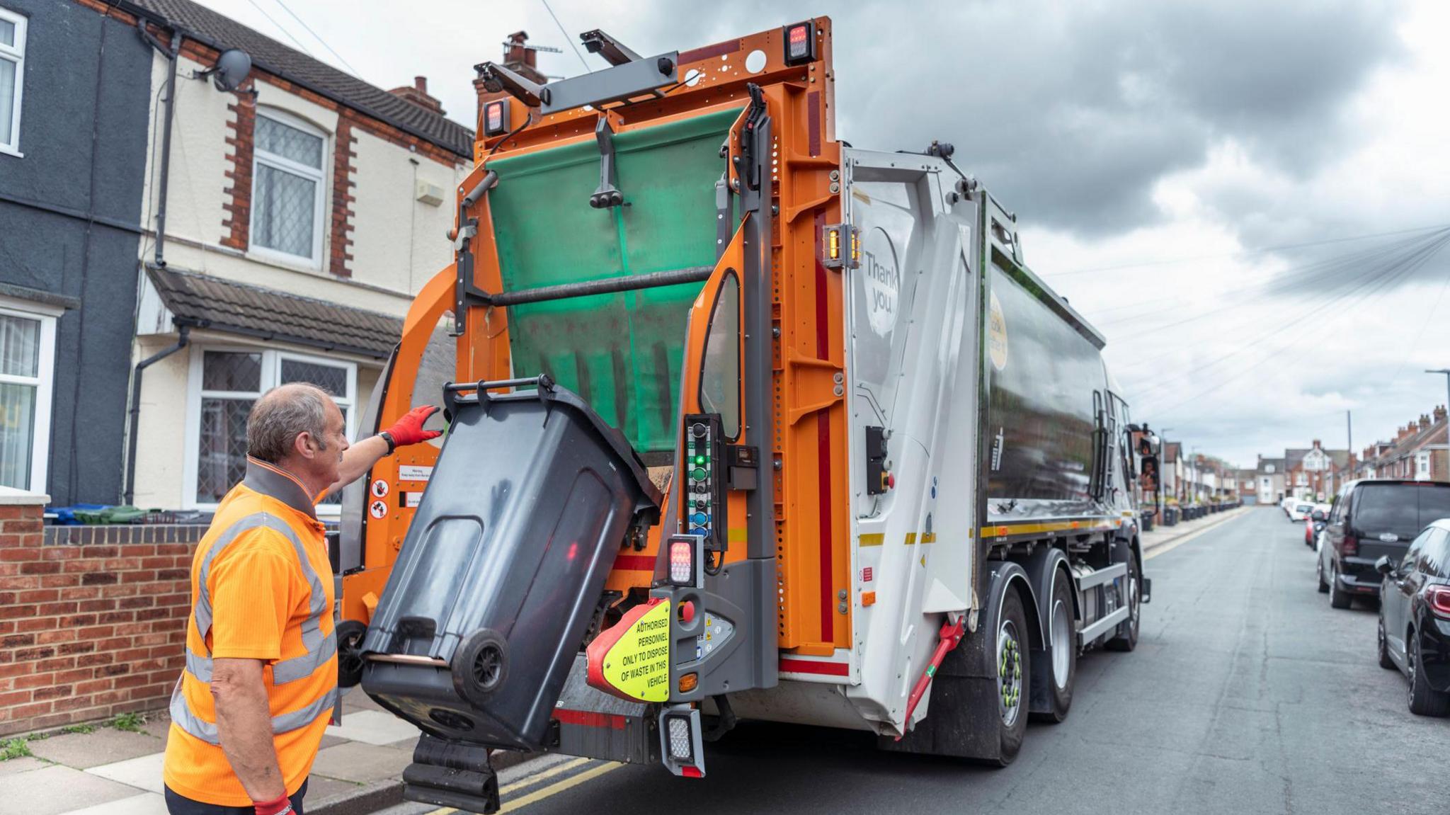 A garbage collector, wearing a bright orange hi-vis vest and red gloves,  emptying a black bin into the back of a large orange and white waste collection truck on a residential street.