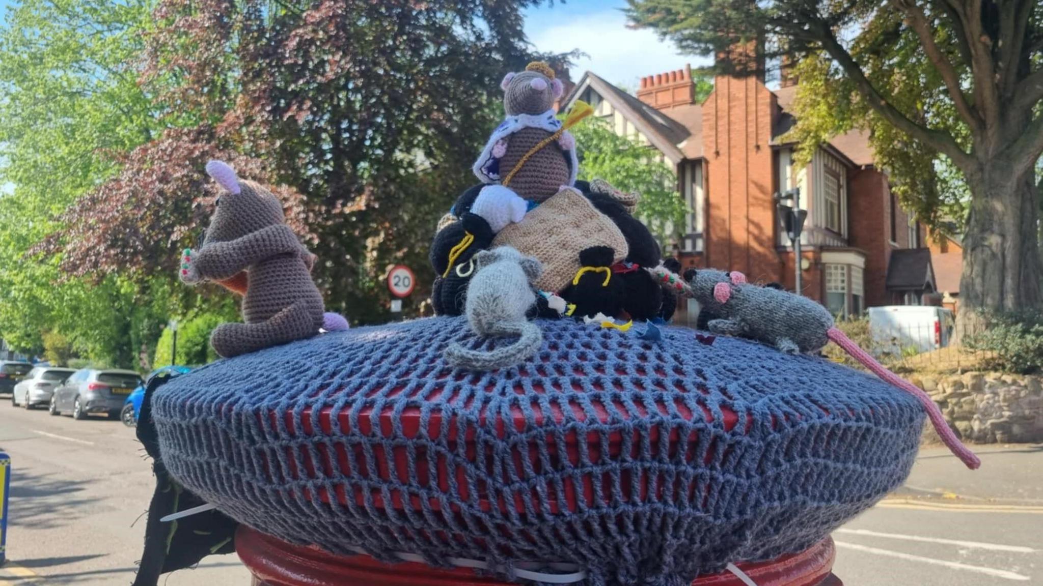A knitted rat, with a crown and a sceptre designed to look like cheese, sits on top of a mound of knitted bin bags on top of a postbox. Other knitted rats can be seen sat around it.