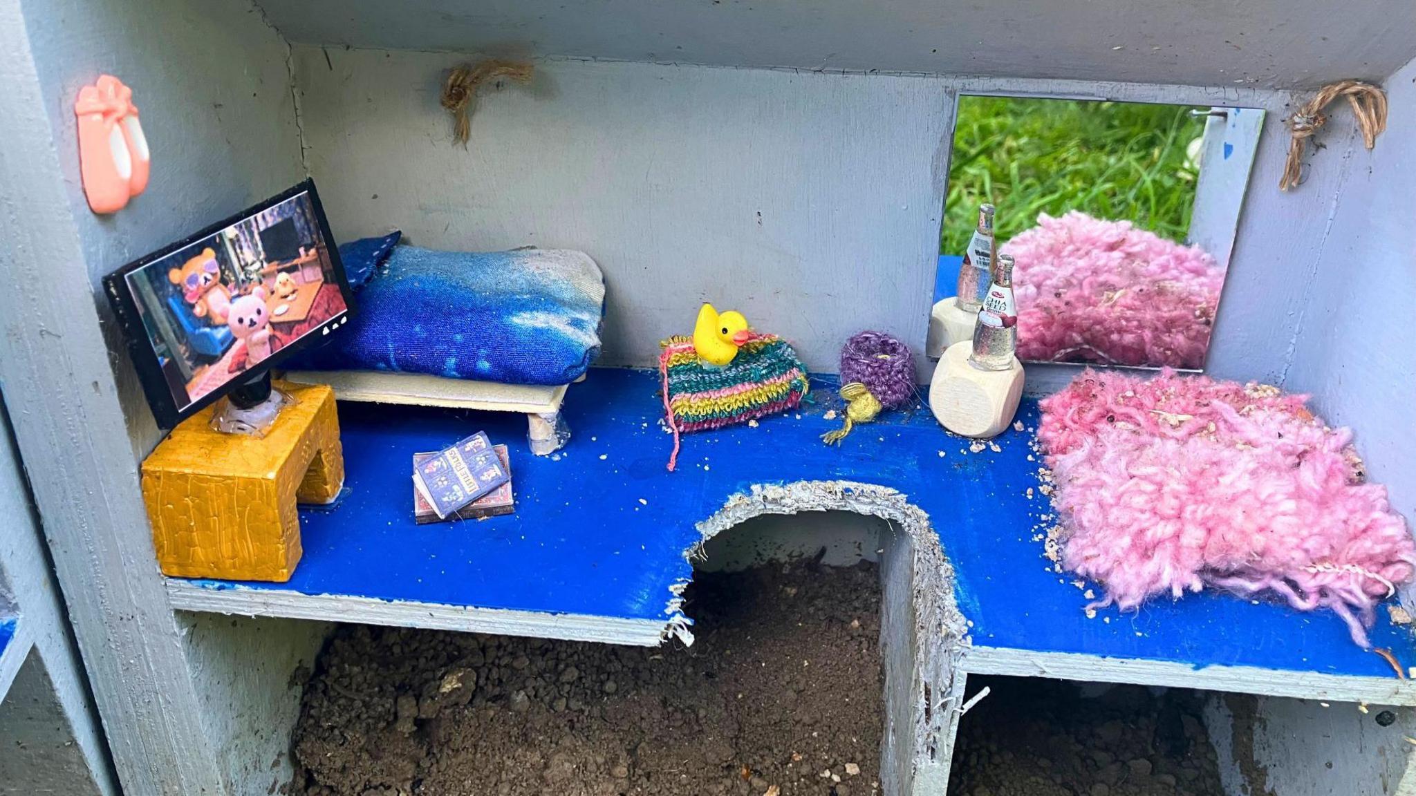 A mini bedroom with a bed, TV, rug and duck is set up in a dollhouse.