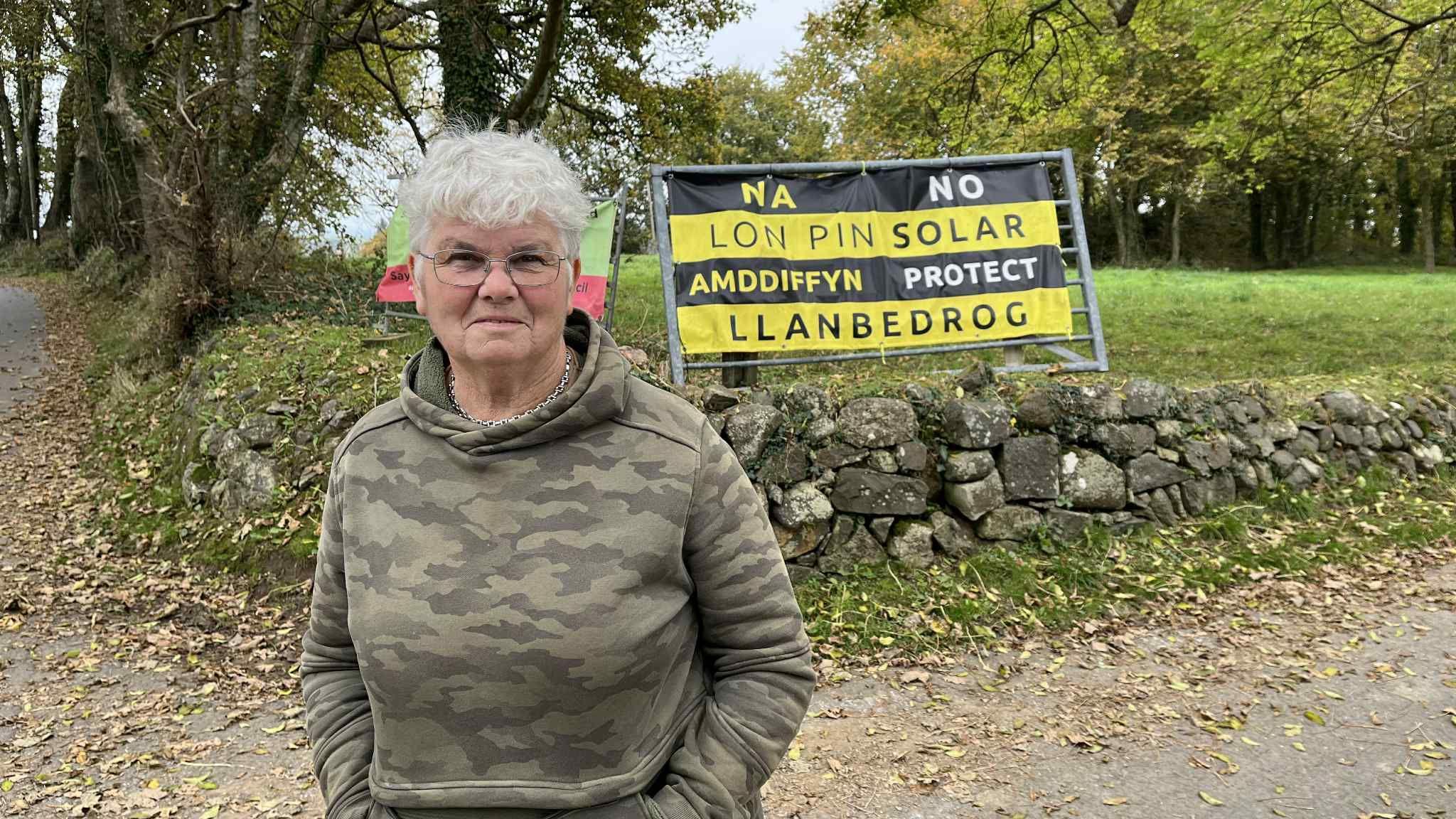 Dr Greta Hughes yn sefyll o flaen baner yn gwrthwynebu'r cais cynllunio