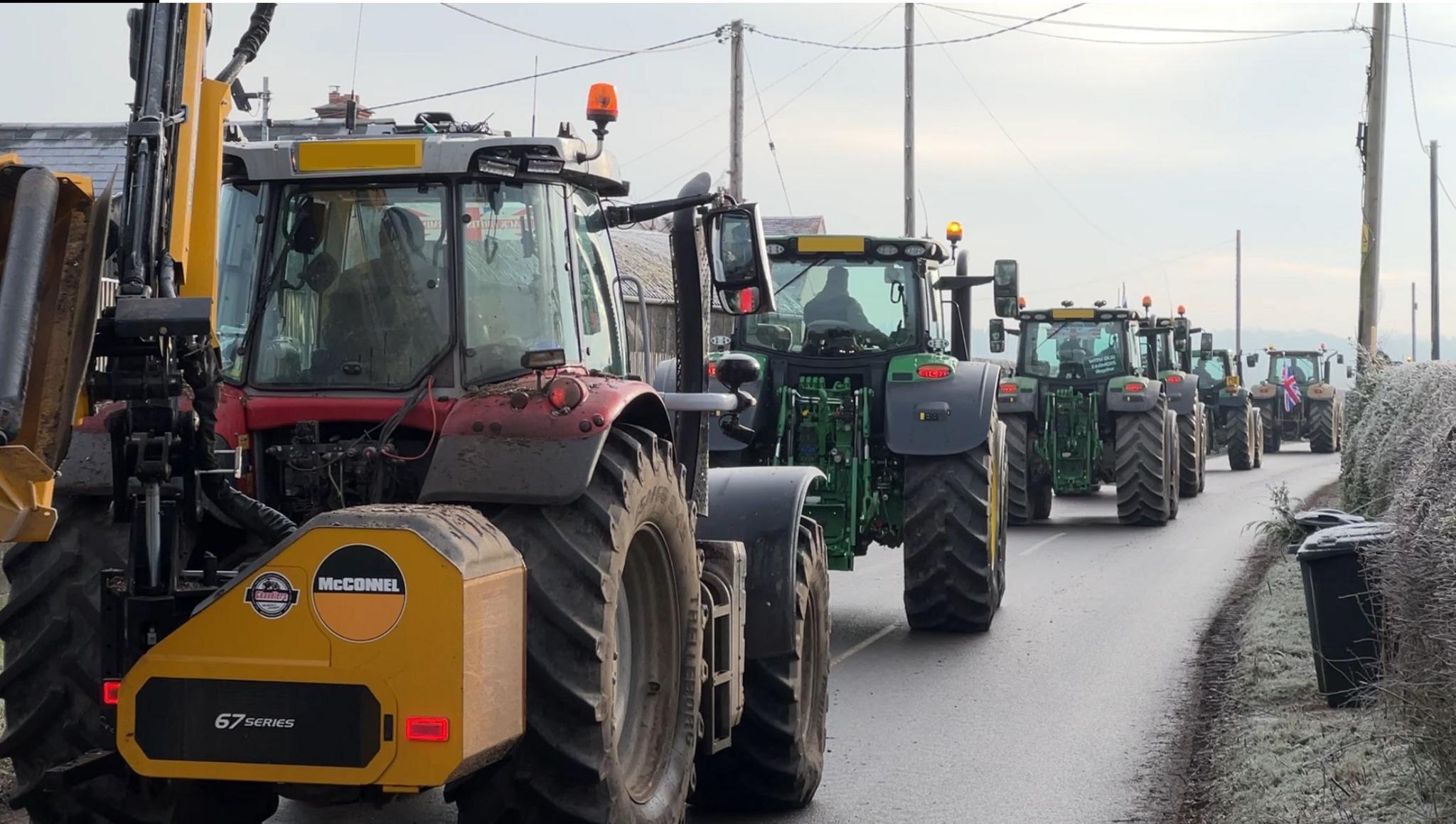 A line of six tractors drive down a rural road. The one nearest to the camera is red and has a yellow piece of farm machinery attached to its rear. The tractors in front are green. The tractor furthest from the camera has a Union Flag attached to its rear. 