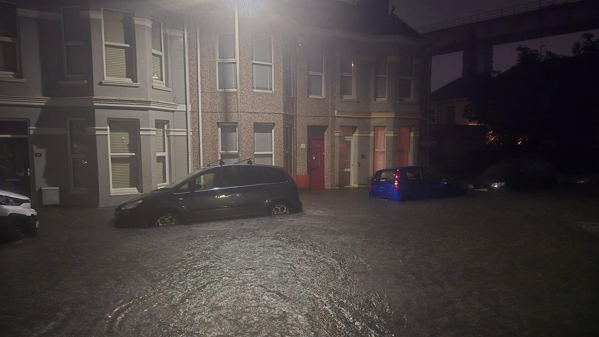 The bottom of parked cars underwater after flooding in Keyham. The picture is taken at night and there are houses across the road.