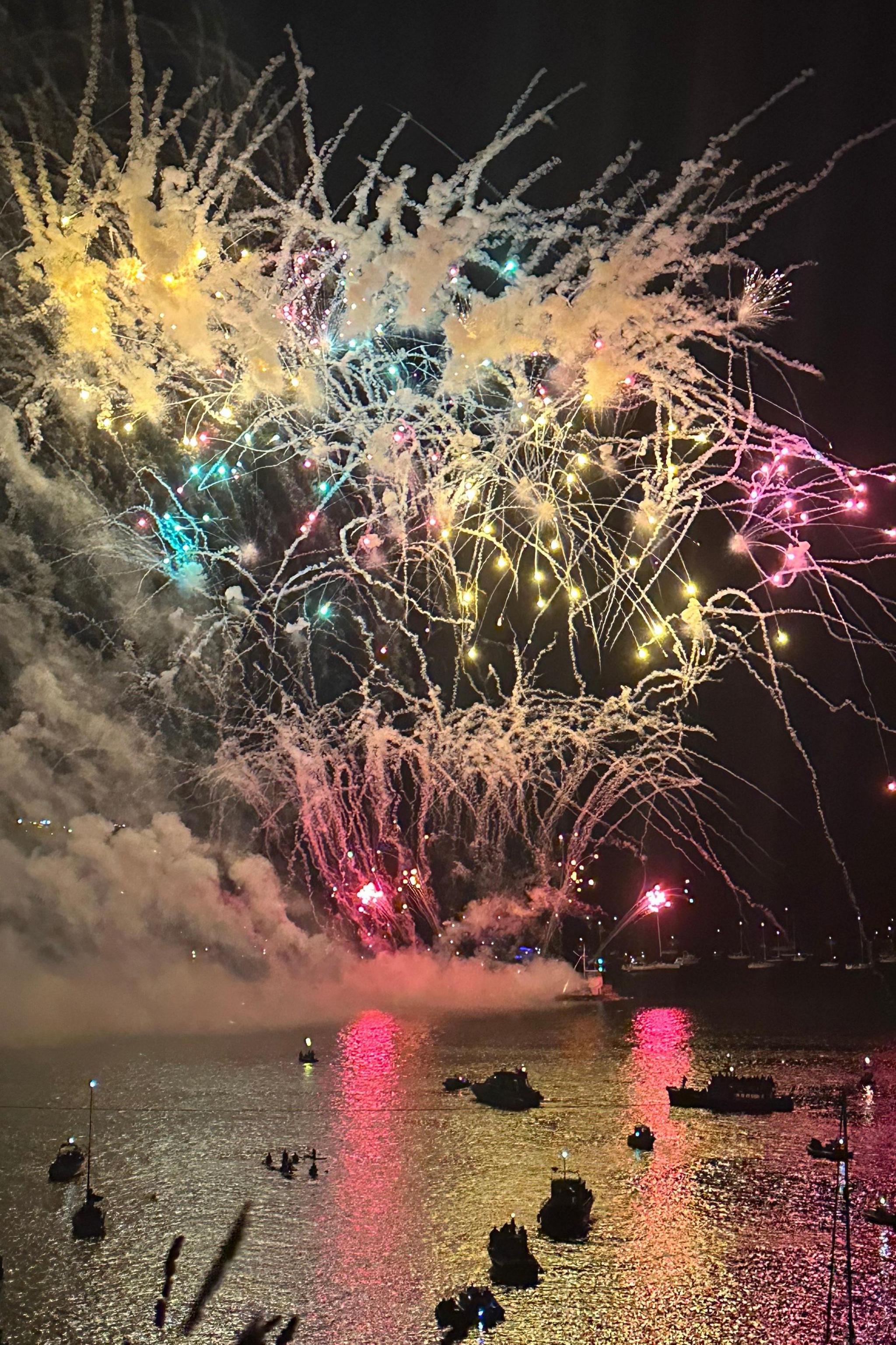 A mass of pink, yellow and blue fireworks full of star shapes and streaming lights exploding above the water. The water is reflecting the colours and has boats of spectators on it.