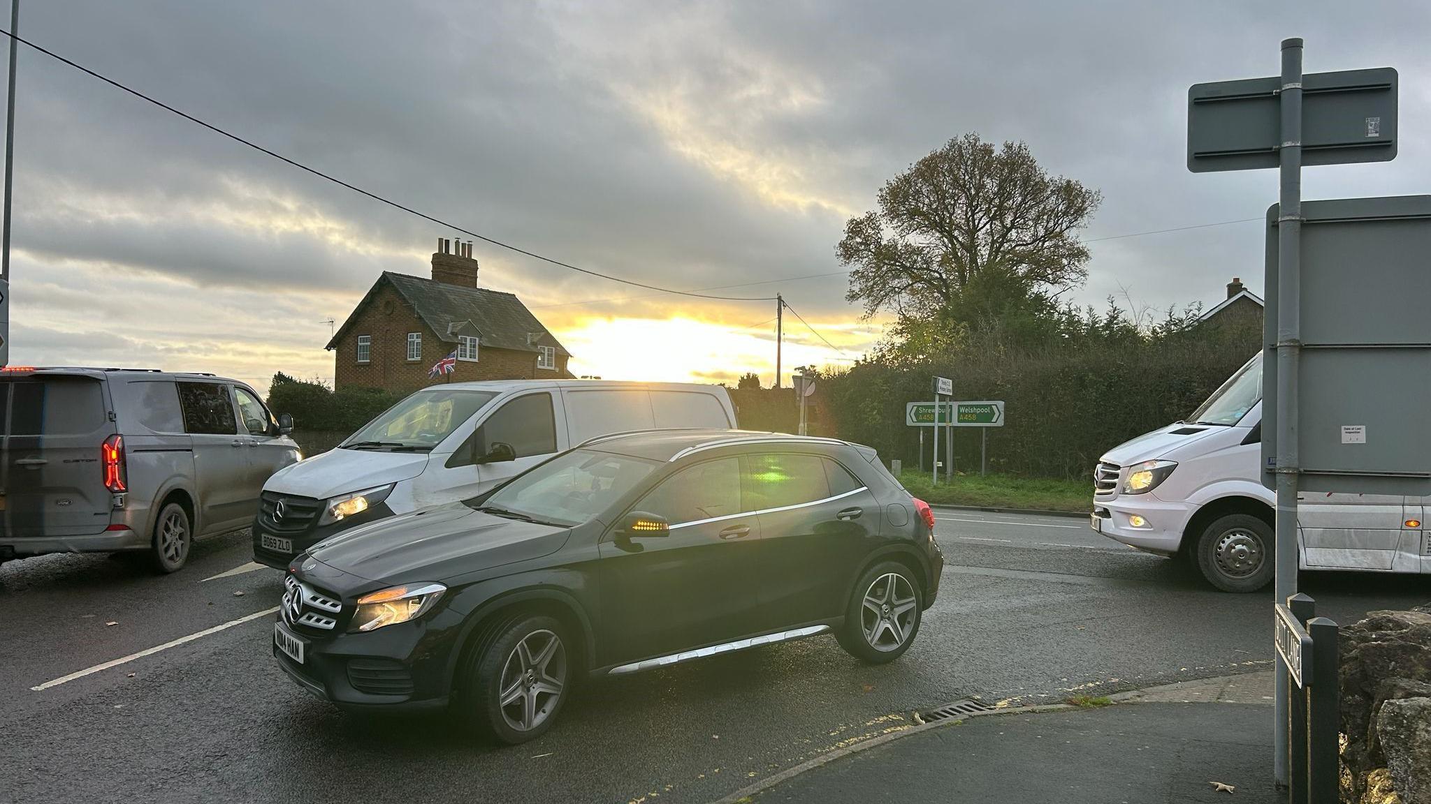 Cars at a busy road junction with the sun rising in the background and a white minibus on the road behind