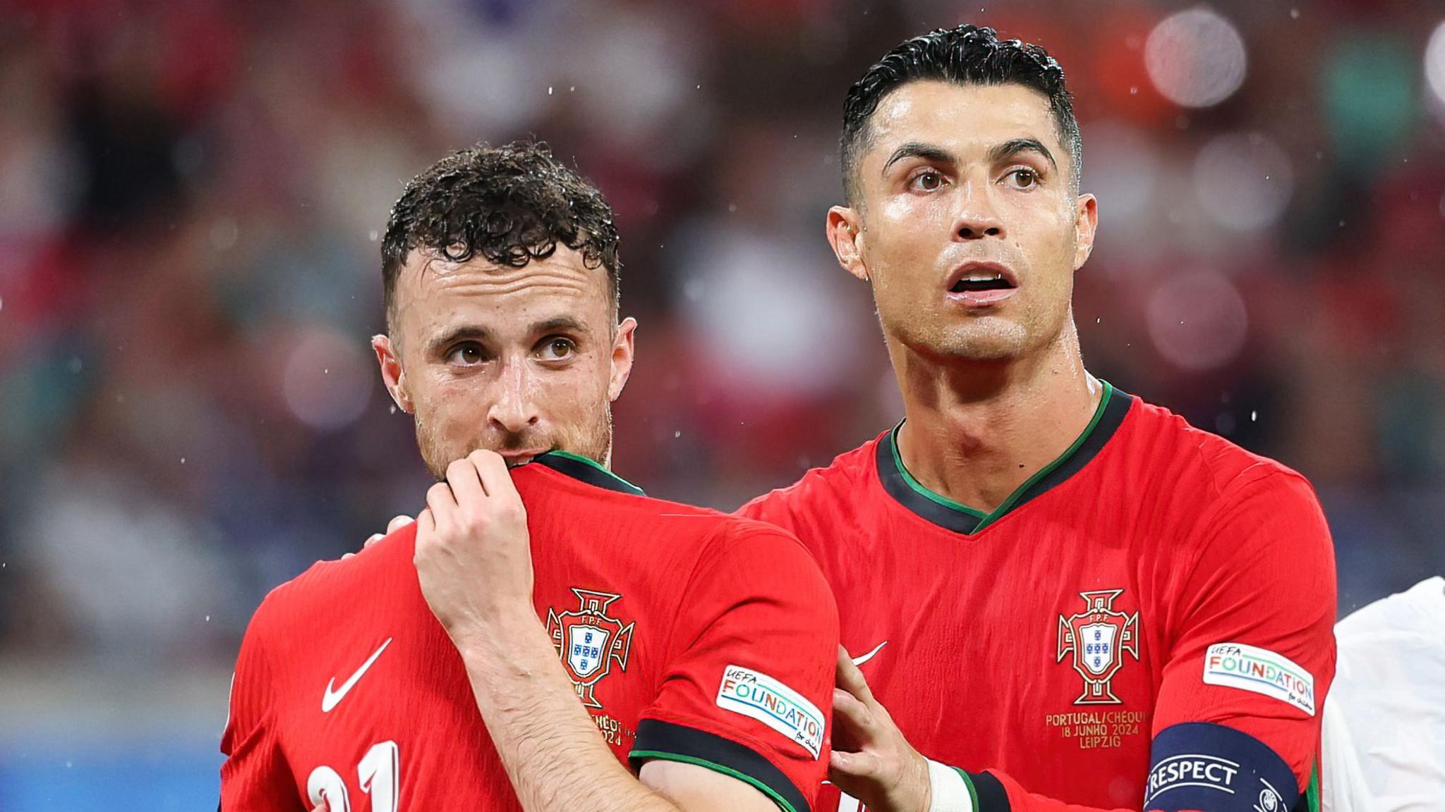 Diogo Jota lifts his shirt to his mouth and looks away from camera while Ronaldo touches his shoulder during a Portugal match