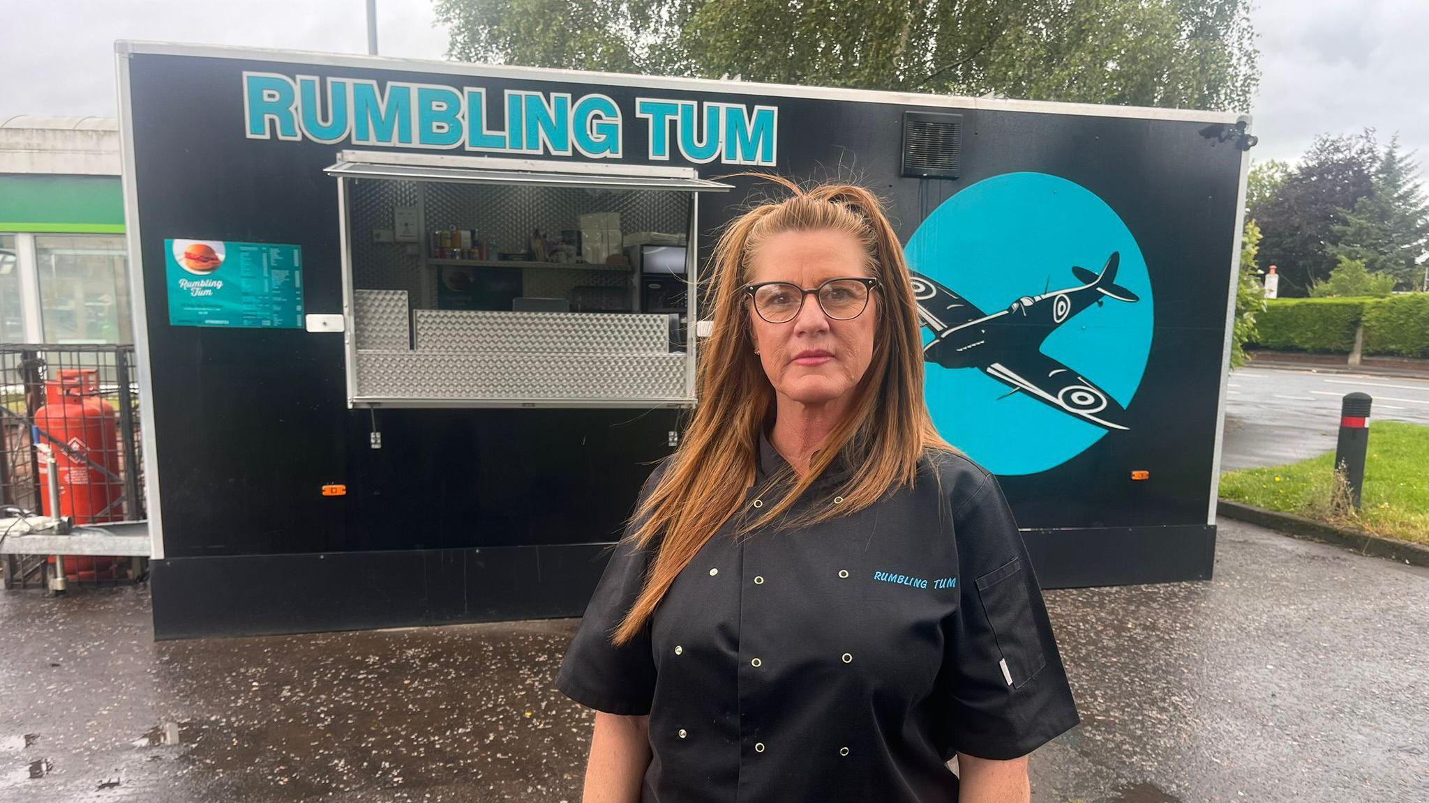 Brenda Bolton, a lady with long chestnut hair held back in a hair band, stands in front of her food van which is painted black and teal with a Spitfire plane on the side and the name "Rumbling Tums".
