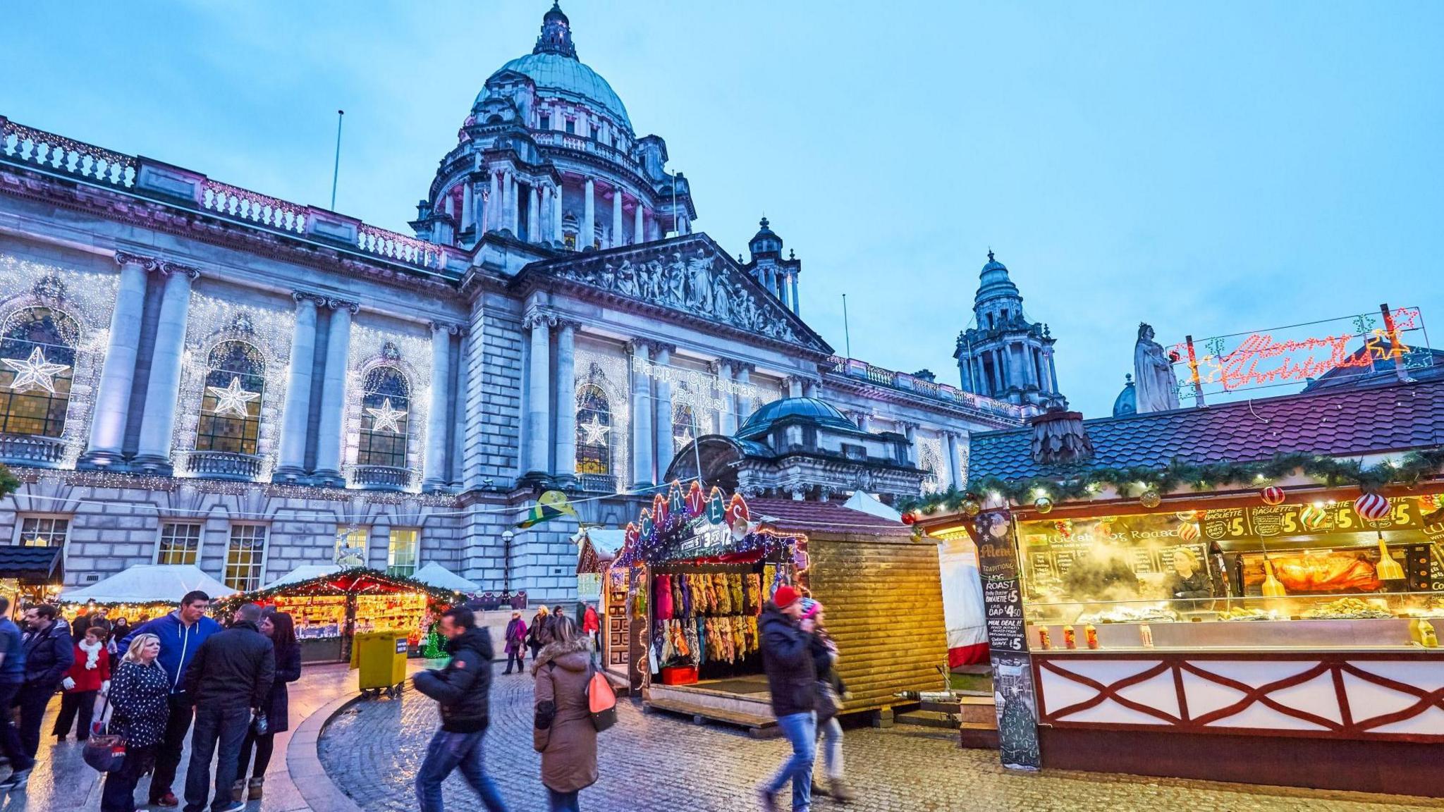 A landscape shot of market stalls on the grounds of Belfast City Hall. There are a number of people walking around.