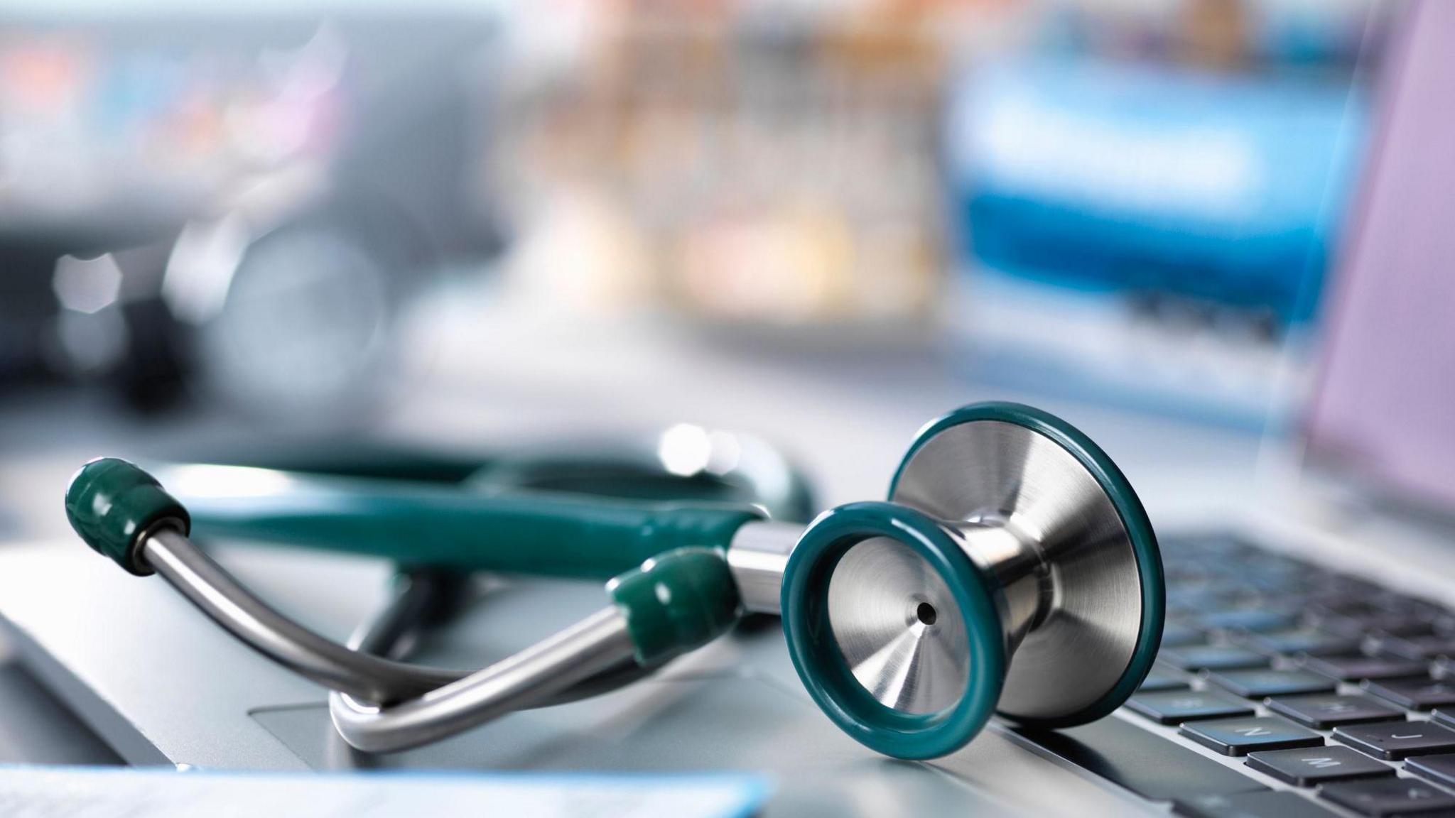 A stethoscope in a turquoise colour with silver metal parts is placed on a laptop trackpad, with the rest of the laptop seen in the background but out of focus.