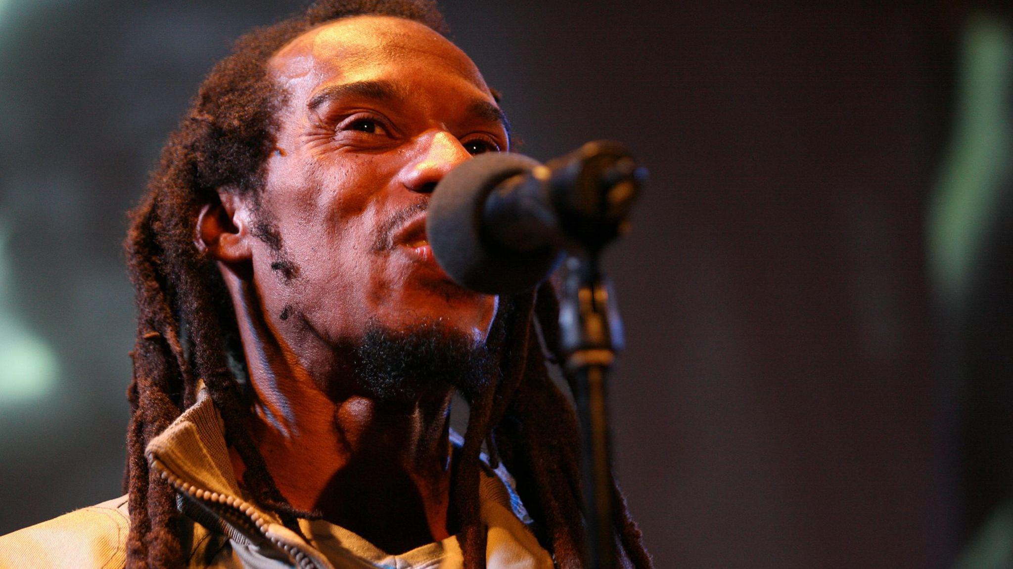 Benjamin Zephaniah speaking into a microphone. He is wearing a beige bomber jacket and has a goatee beard and dreadlocks.  