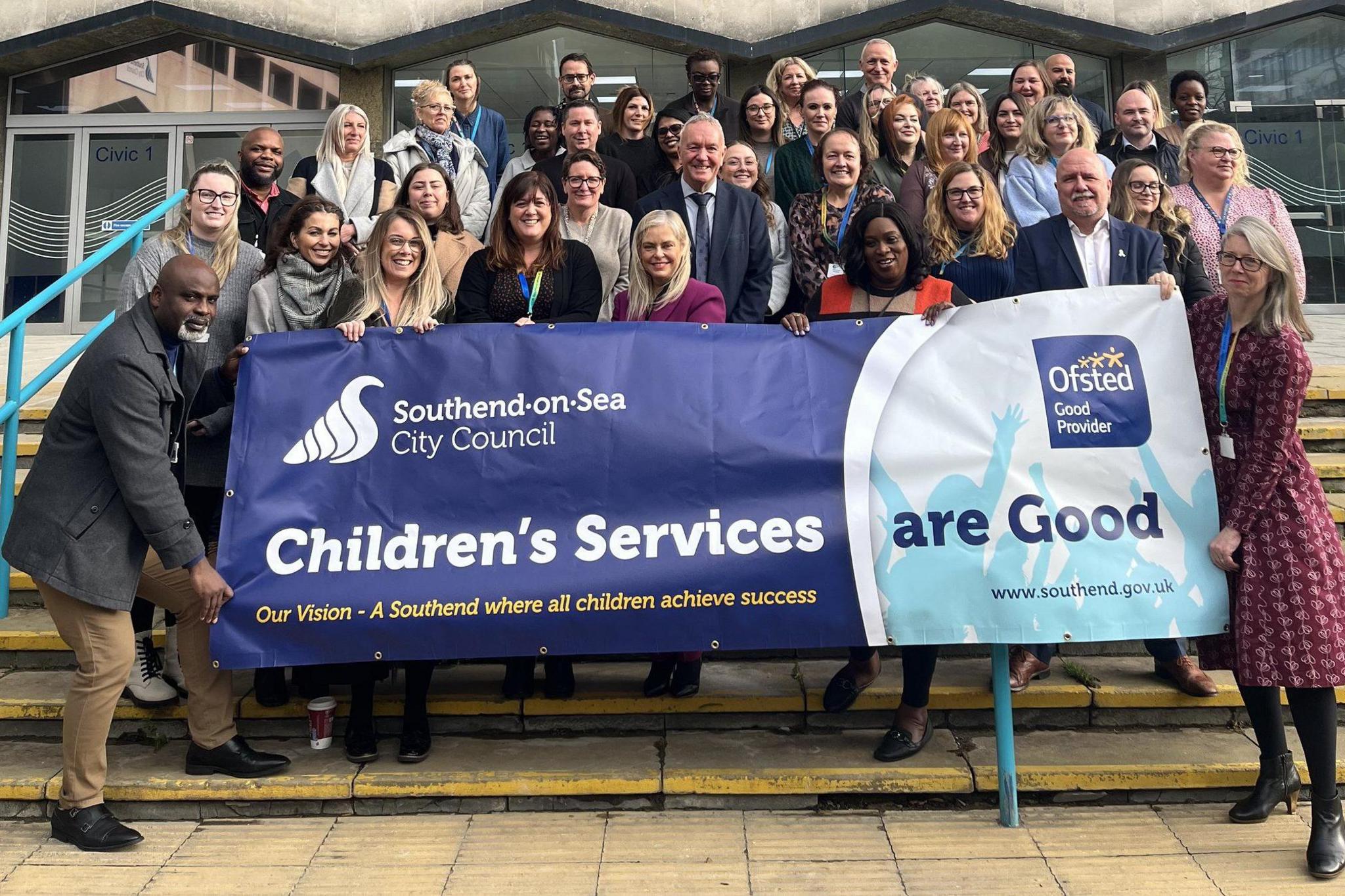 About 40 people are standing on steps outside Southend-on-Sea City Council's offices.  The people at the front are holding a banner, which reads "children's services are good" with the Ofsted logo on it.