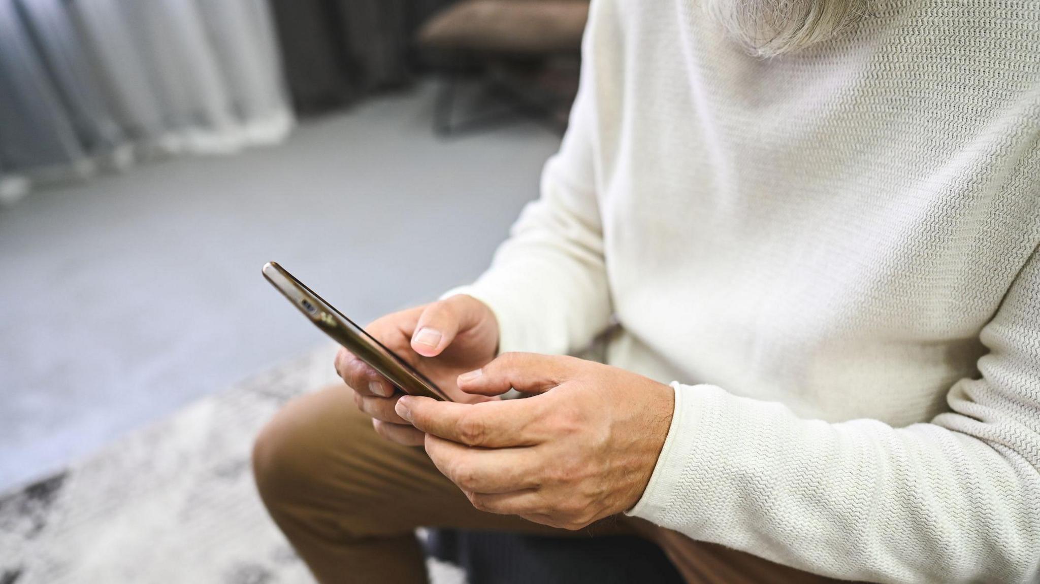 A close up on a man's hands using a mobile phone. He is wearing a cream jumper and brown trousers and sitting in a room with a grey rug and carpet.