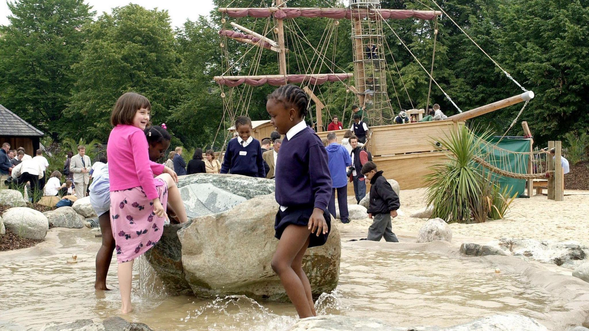 Children playing around a rock in a shallow pool. A play galleon is resting in sand behind them