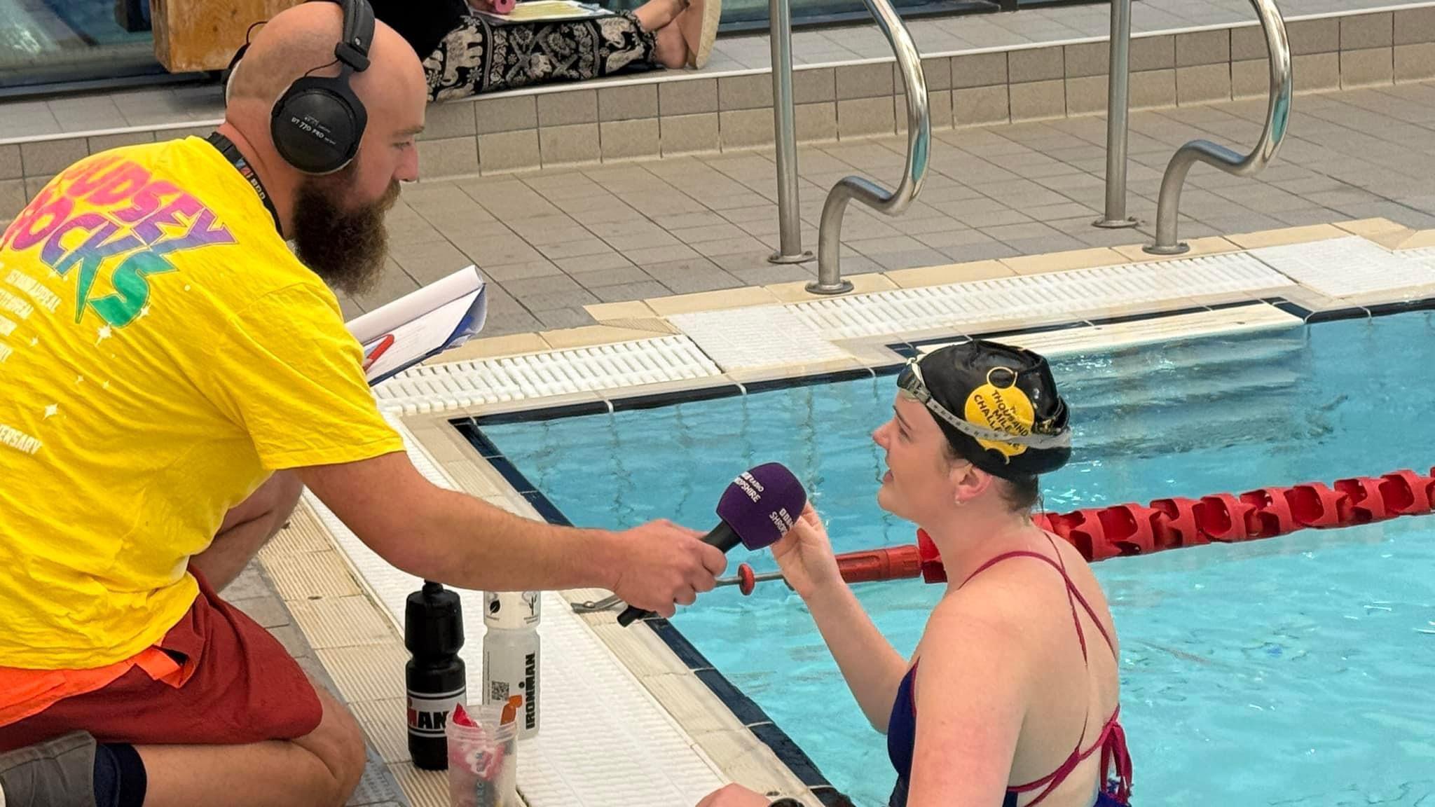 Charlotte is standing in a swimming pool wearing a black Children in Need swimming cap. She is speaking into a purple microphone. A man with a bald head and long beard is wearing a yellow Children in Need T-shirt and is crouching on the side of the pool, holding the microphone to Charlotte's mouth.