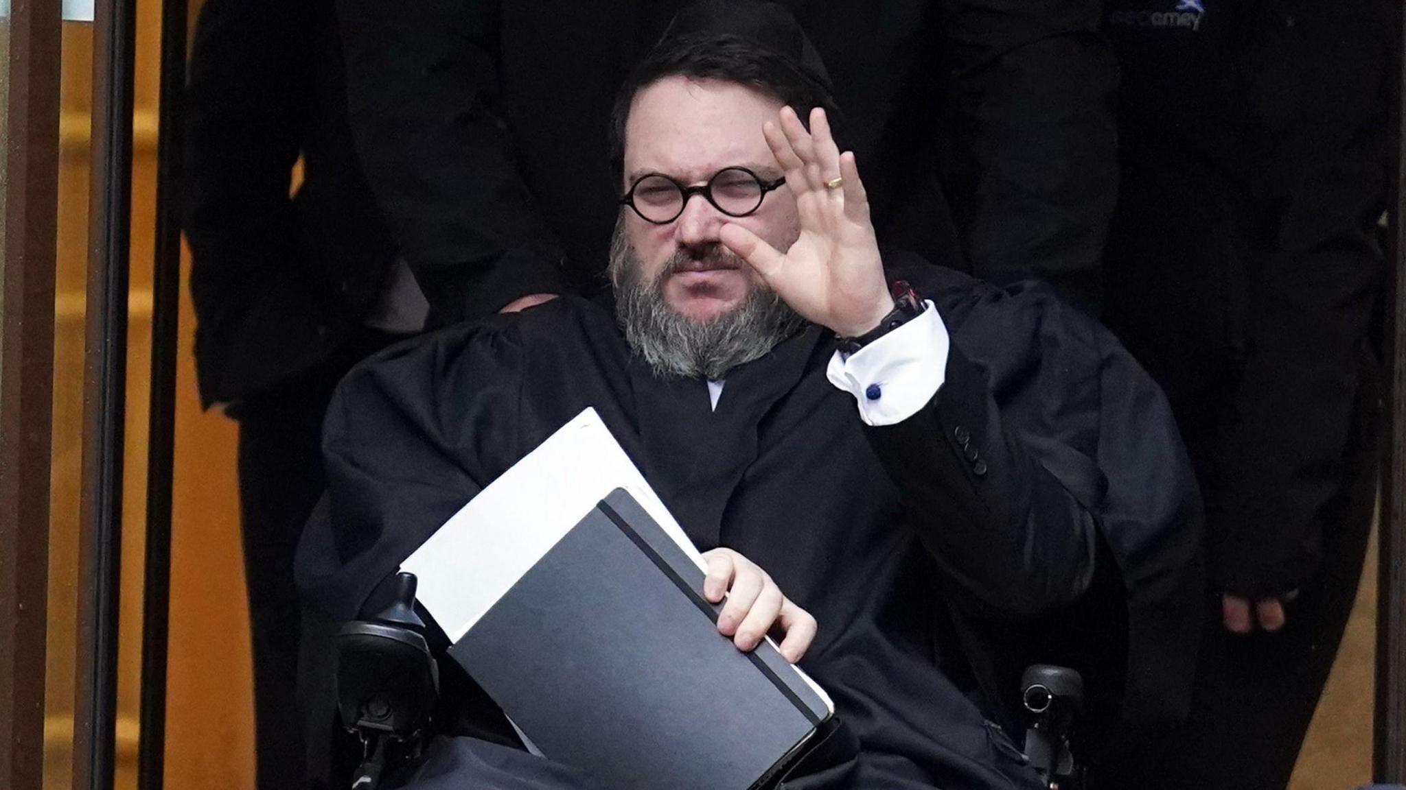 Nicholas Rossi wearing all black. He has a white cuff on his left arm, which is raised in front of his face. His right arm is clutching a black notebook with a white A4 page sticking out of it. He is seated and has a grey-black beard and dark rimmed glasses on. He has dark hair pushed to the left.