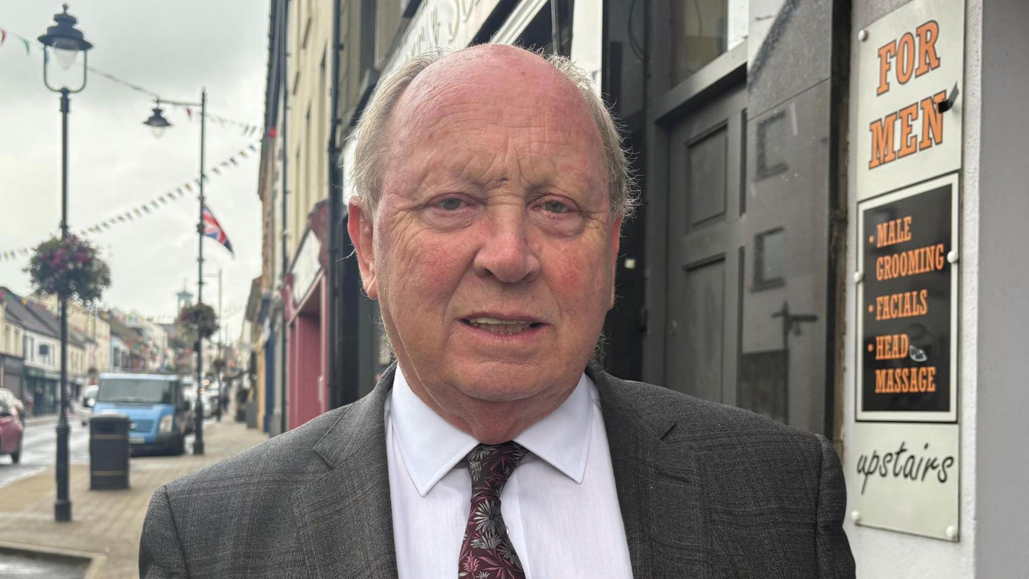 A man wearing a grey suit, white shirt and floral tie looks to the camera. He has grey hair on the back and sides of his head. He is standing in the street of a town. Vehicles can be seen on the road in the background, as can lampposts, flowers, red, white and blue bunting and a union flag.