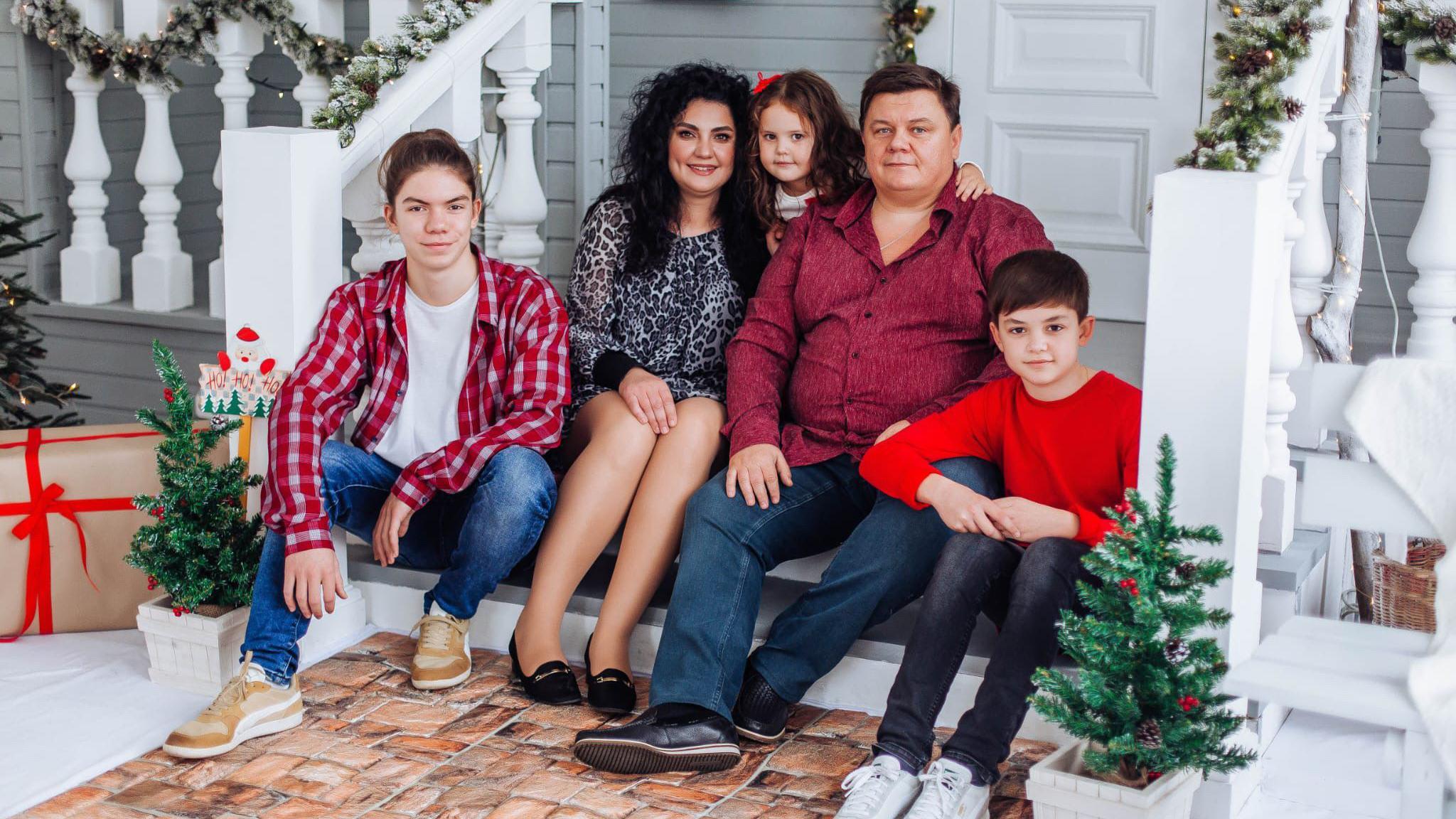 A portrait of Nataliya Korovina's family. They are sitting in front of a white house with Christmas trees, presents and wreaths on its wooden porch. On the left is the family' oldest son, Nataliya is next to him. She has her arm around their daughter. Nataliya's husband has short brown hair and is wearing a red shirt. Below him is their youngest son, who has short brown hair.