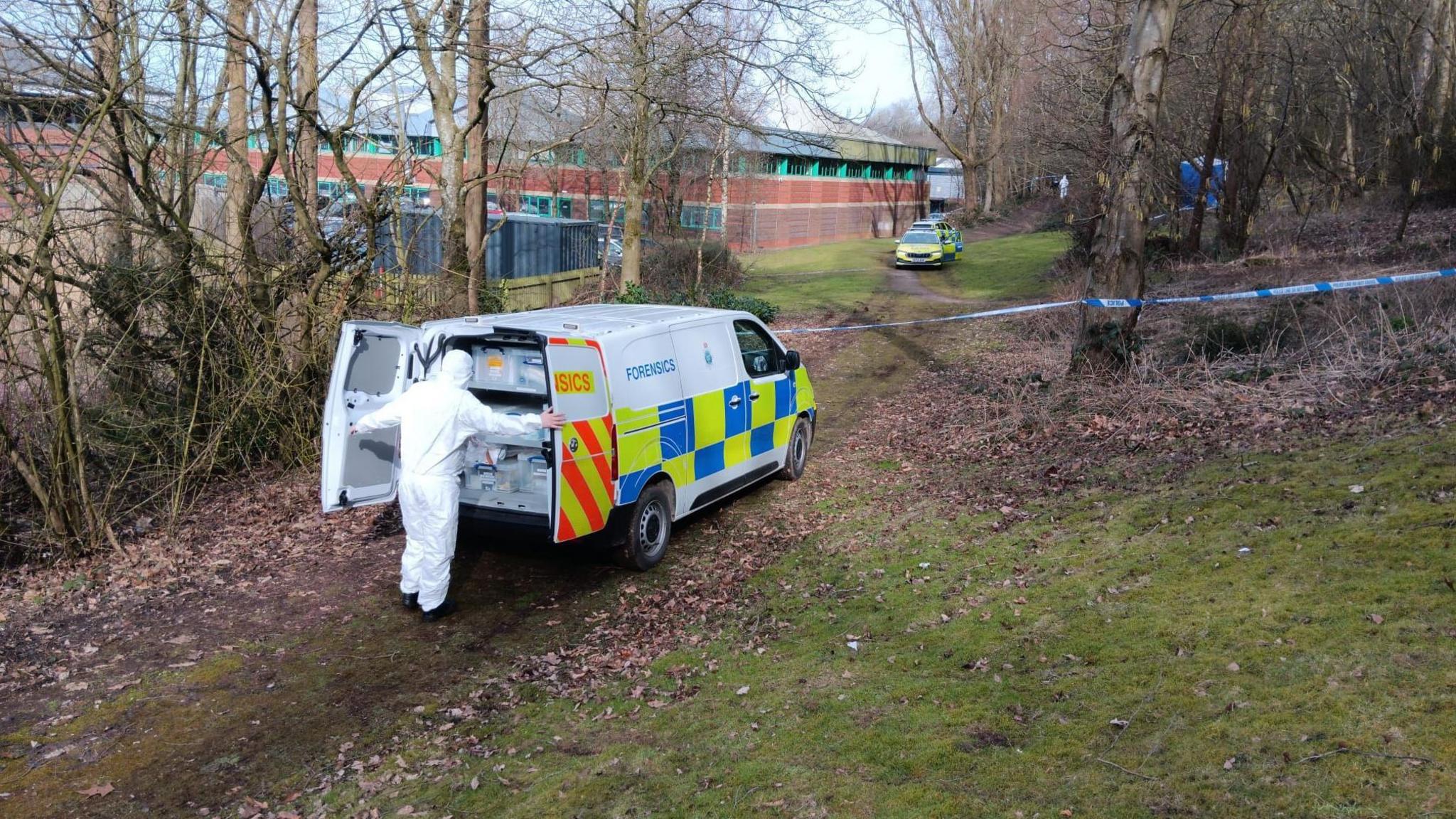 A forensics officer at the back of a police vehicle. They are wearing a white protective suit and are looking inside the vehicle which says Forensics on the side. In the distance is a blue tent, a police tape and a police car and a building. 