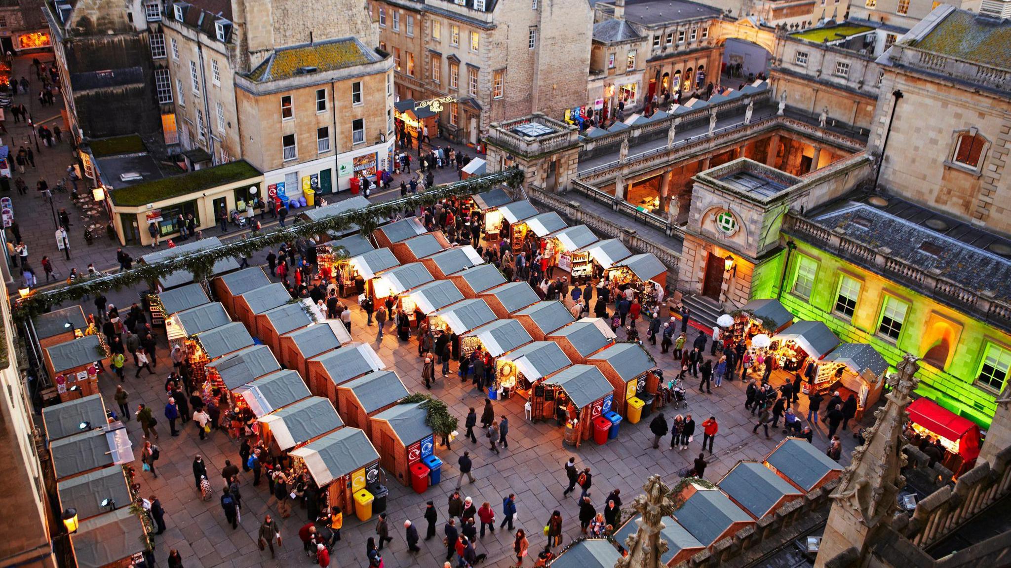 Bath Christmas Market at dusk. There are about 40 stalls with grey roofs lined up in rows. They are lit up with orange Christmas lights and there are lots of people browsing in the market square