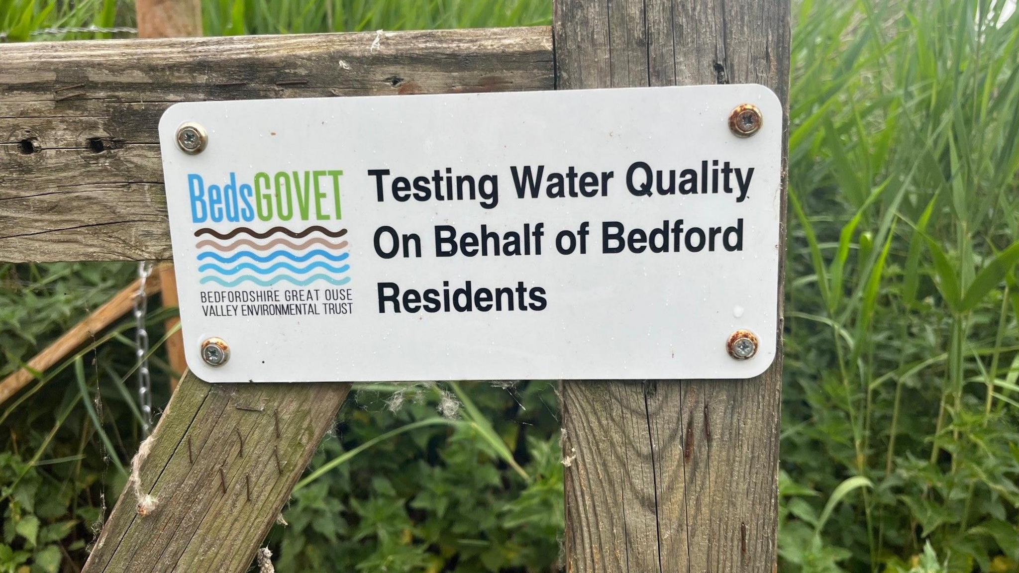 A close up shot of a small white sign that is nailed to a wooden gate. It has the BedsGOVET logo in blue and white, with five wiggly lines below the word in different shades of brown and blue. In black font, the sign says "Testing Water Quality on Behalf of Bedford Residents".