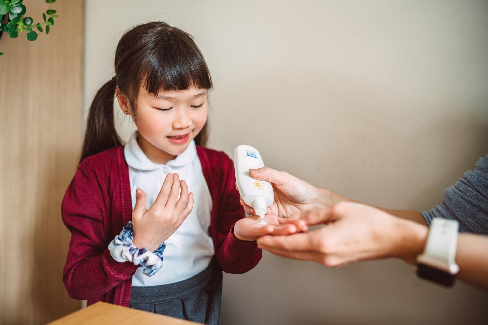 Young Asian girl in school uniform applying sunscreen lotion to her hands before heading out for school, assisted by her parent. 