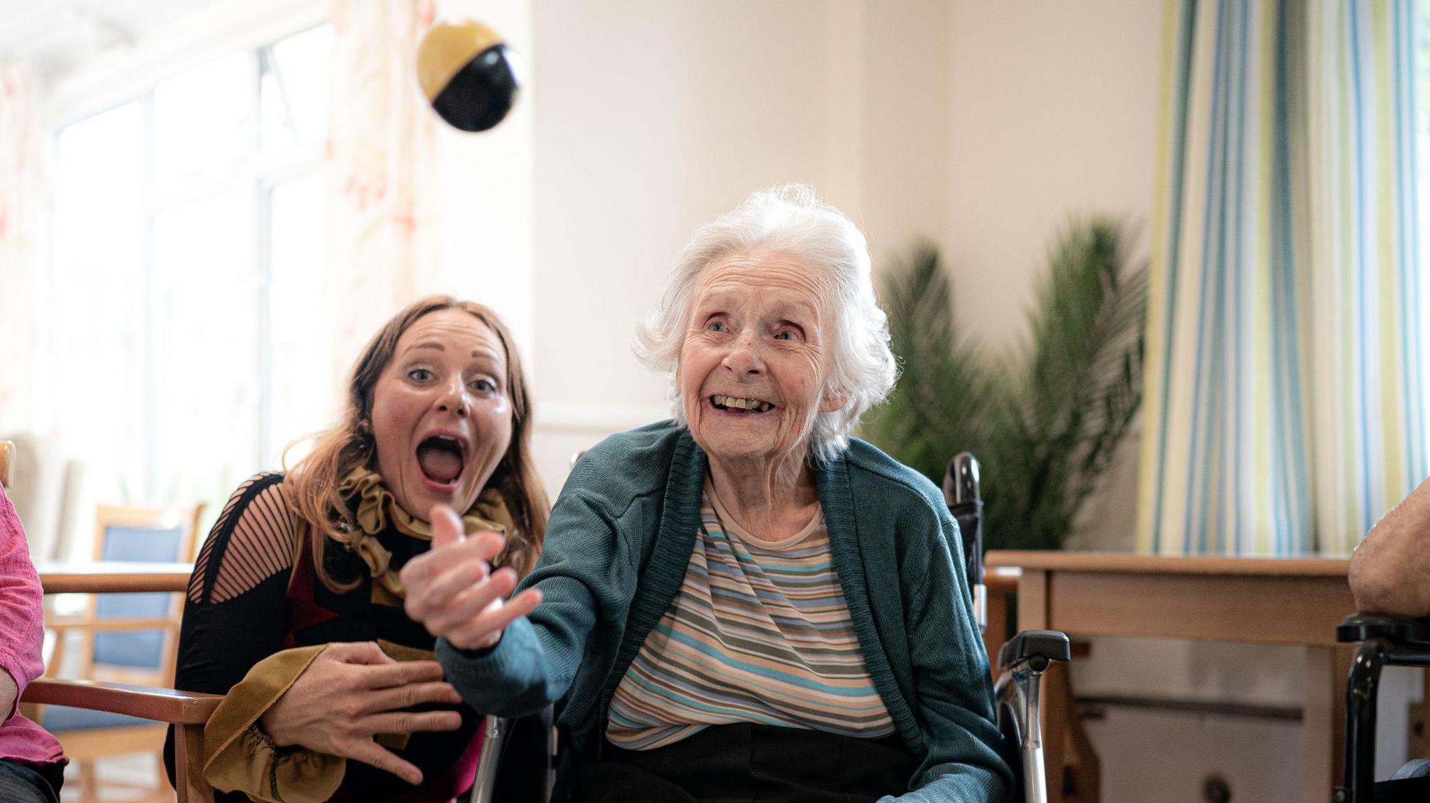 An elderly woman throwing a ball in the air, with a younger woman smiling behind her.