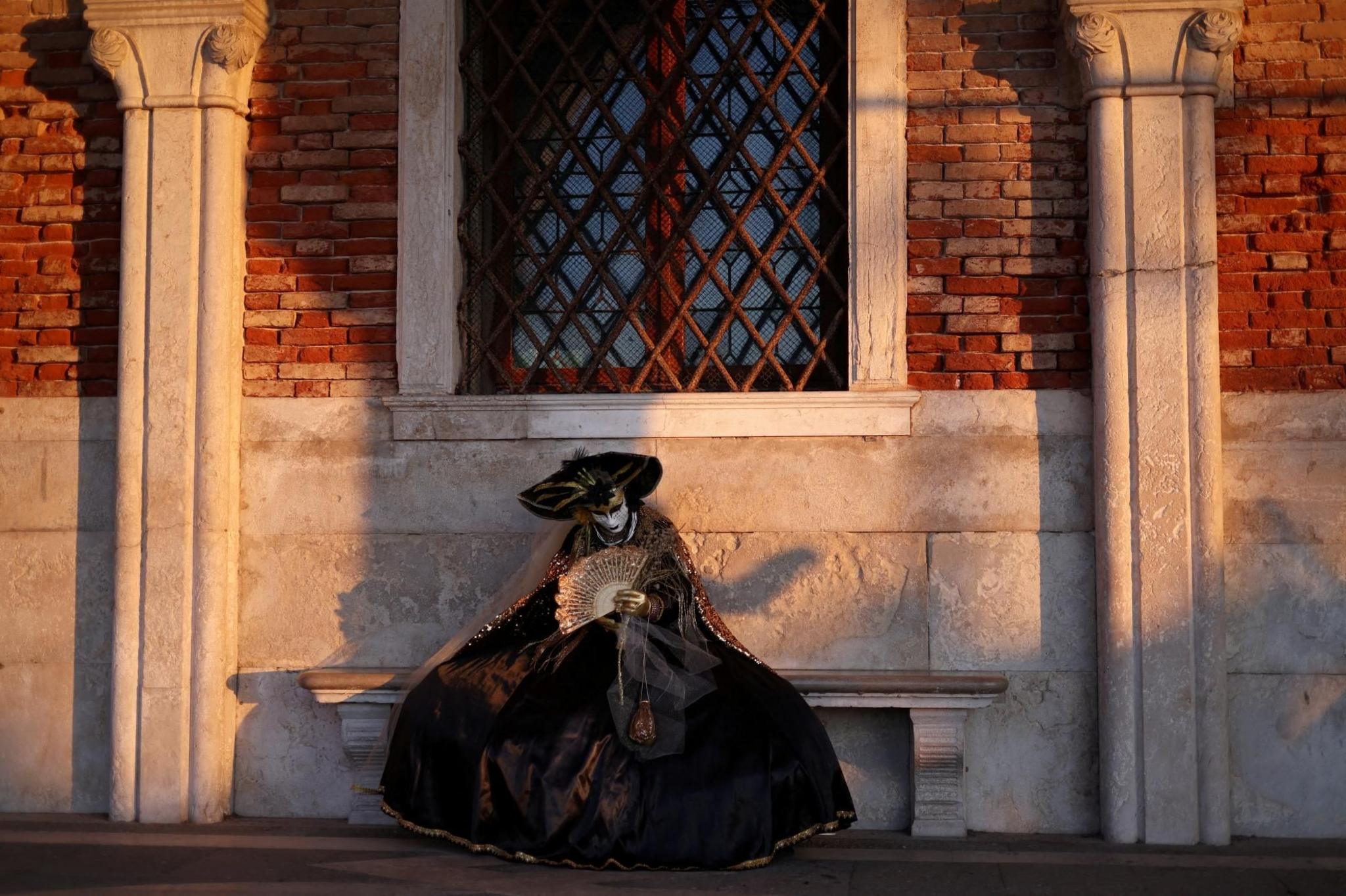 A masked reveller sits alone on a stone bench beside a brick wall. The reveller is wearing a long, dark-coloured cloak, gown and a wide-brimmed hat and is holding a gold-coloured fan.