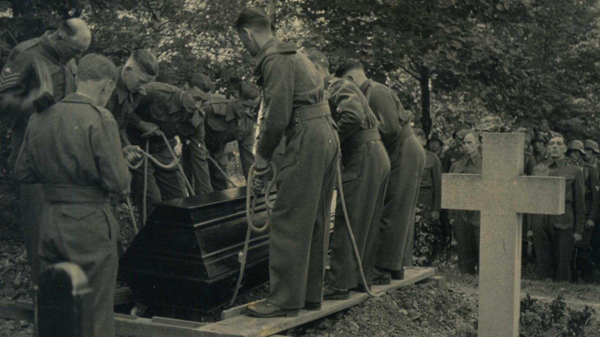 A monochrome photograph from the 1940s which shows a burial ceremony in Berlin. Soldiers are lowering a wooden casket into a grave using a rope system. They stand on wooden boards beside the grave as they lower the coffin. A large stone cross is seen on the right-hand foreground.
