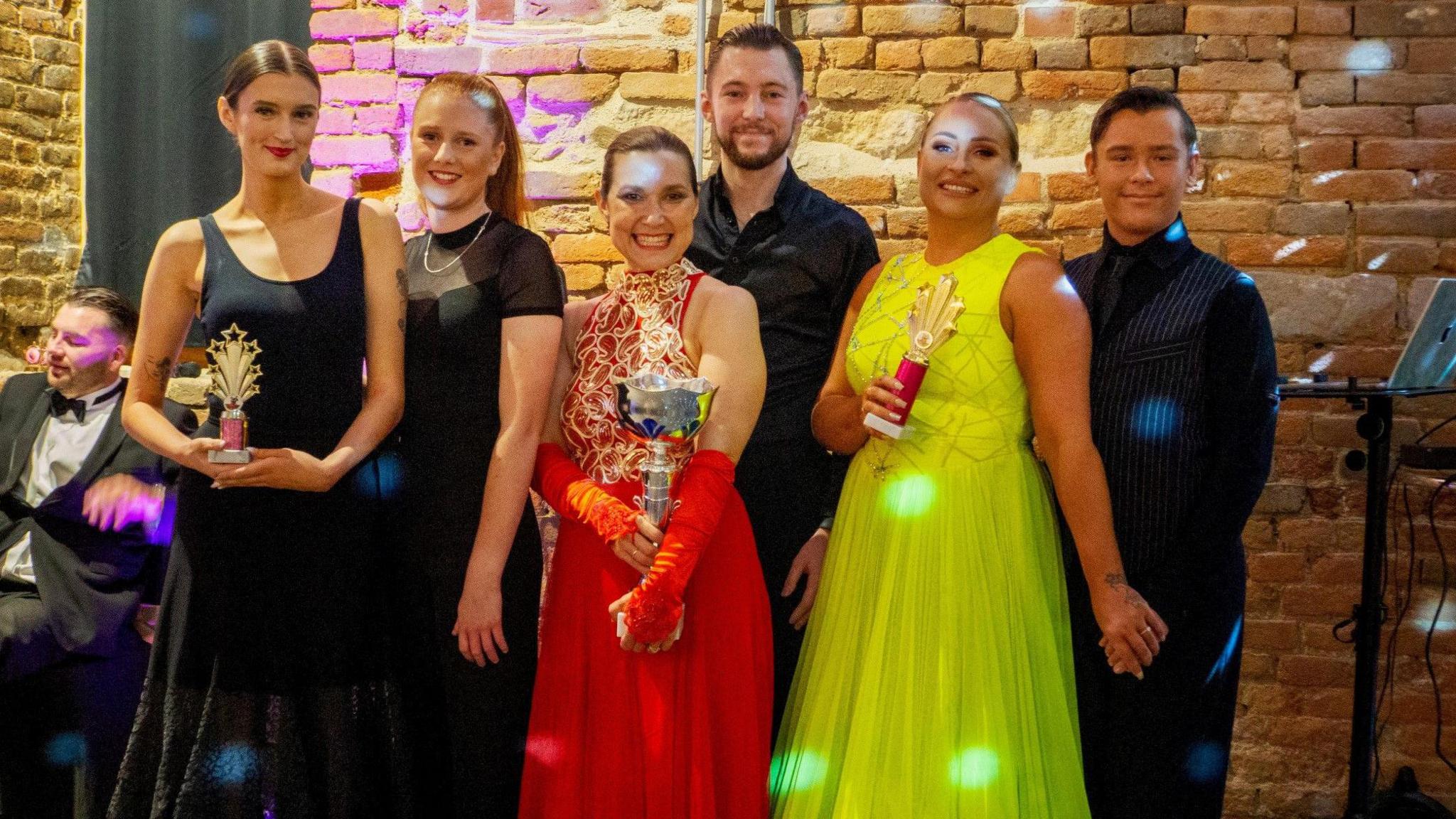 Six people wearing evening wear smile at the camera. Three of the women hold trophies in their hands