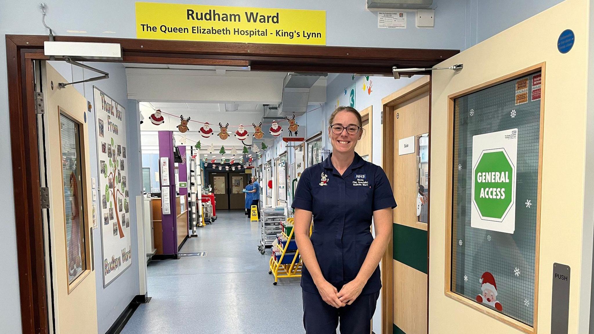 Nicola Wright stands below a sign which says Rudham Ward. All over the walls and doors are Christmas decorations and garlands with pictures of Father Christmas and reindeer. 