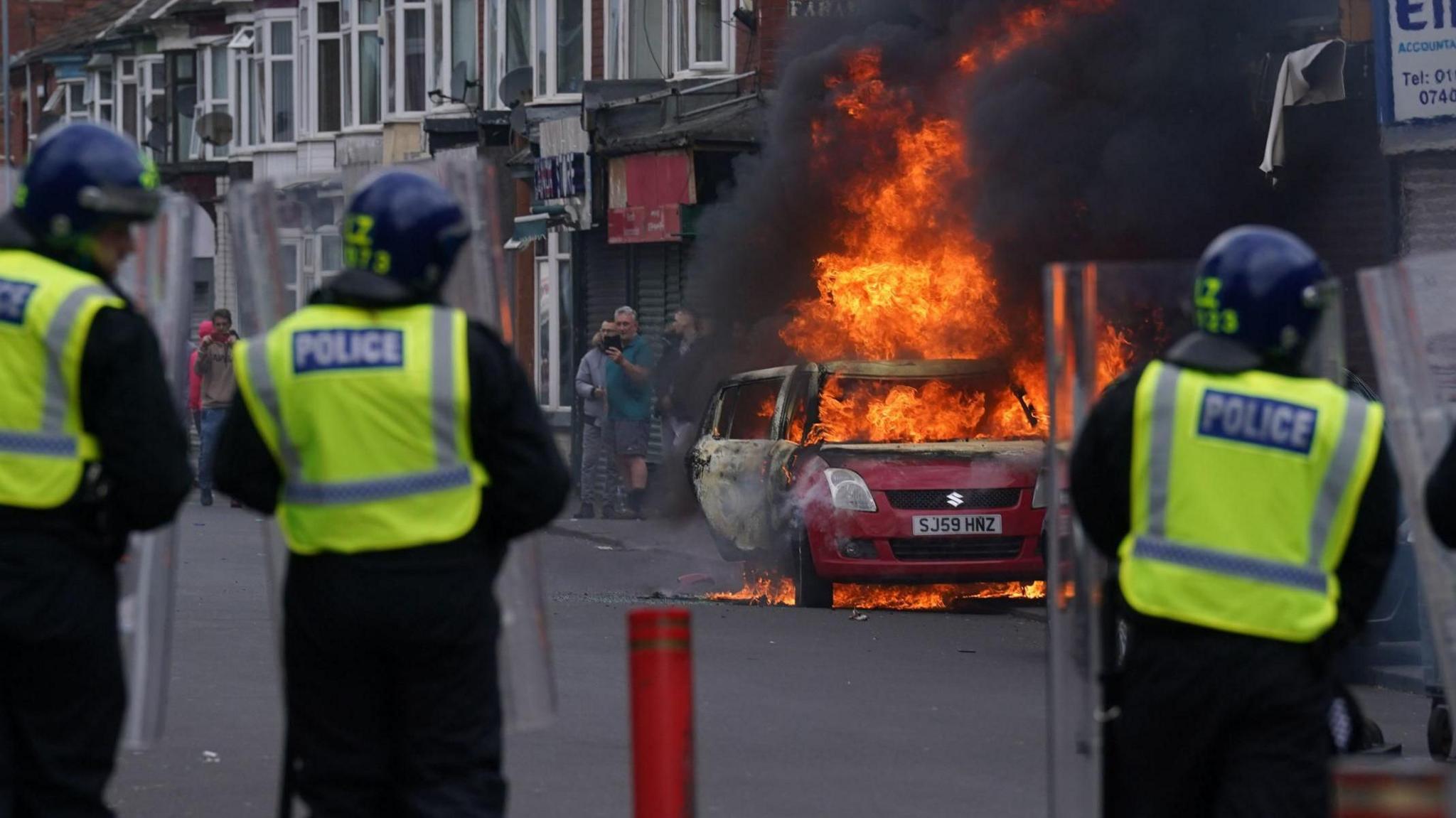 The backs of three police officers are visible as they stand a safe distance from a burning red car on a street. A row of shops can be seen in the distance. A group of people stand beyond the blazing car.