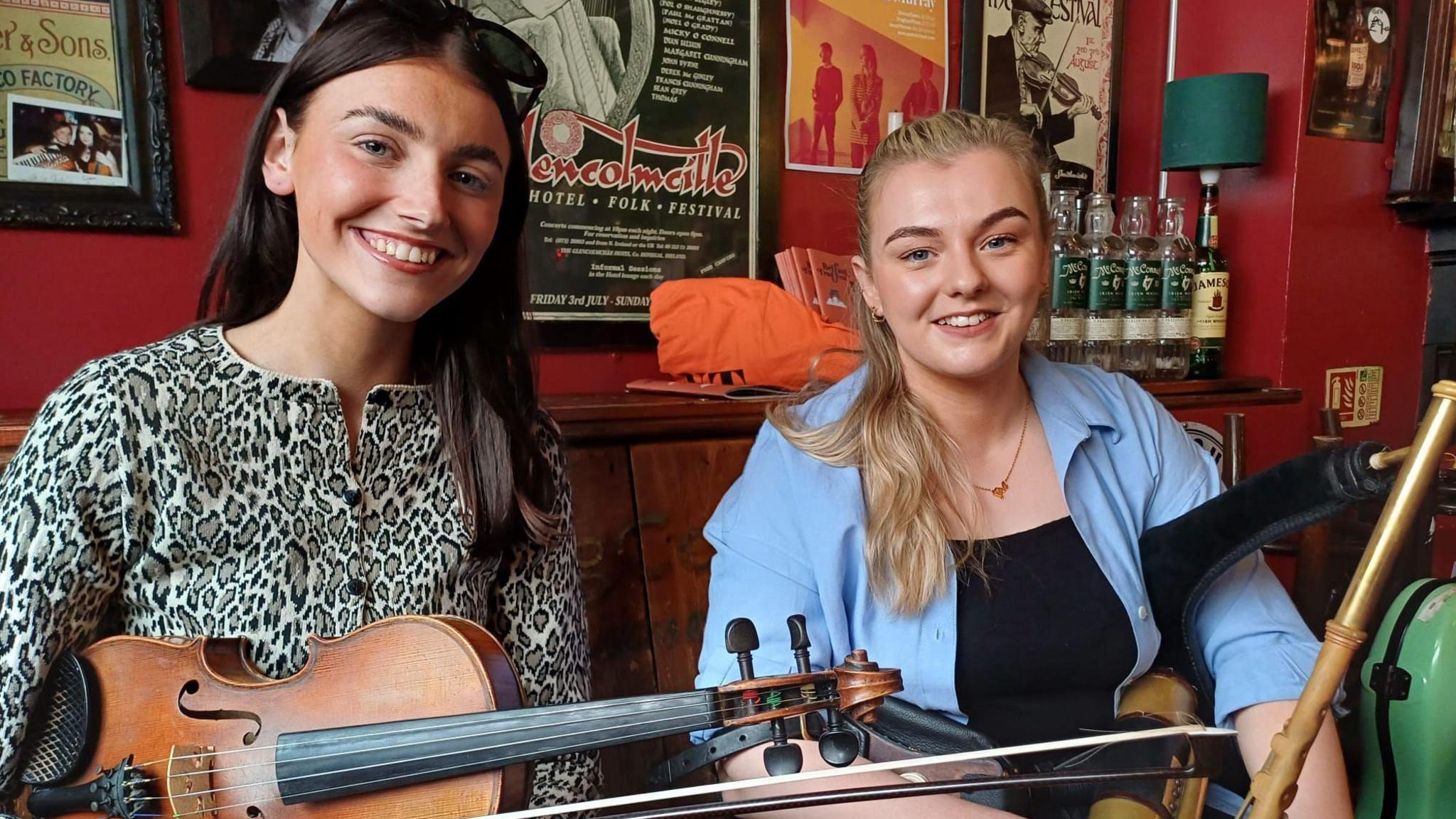 Rosie is smiling at the camera holding her fiddle and Maeve is sitting next to her, smiling at the camera holding her pipes