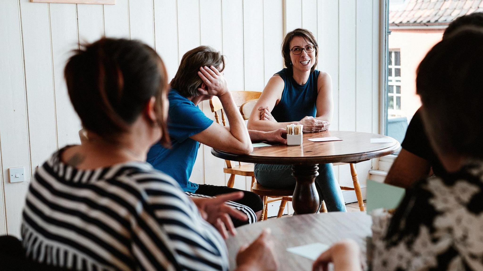 A group of people at a coaching session, in a room with a table, and talking to each other