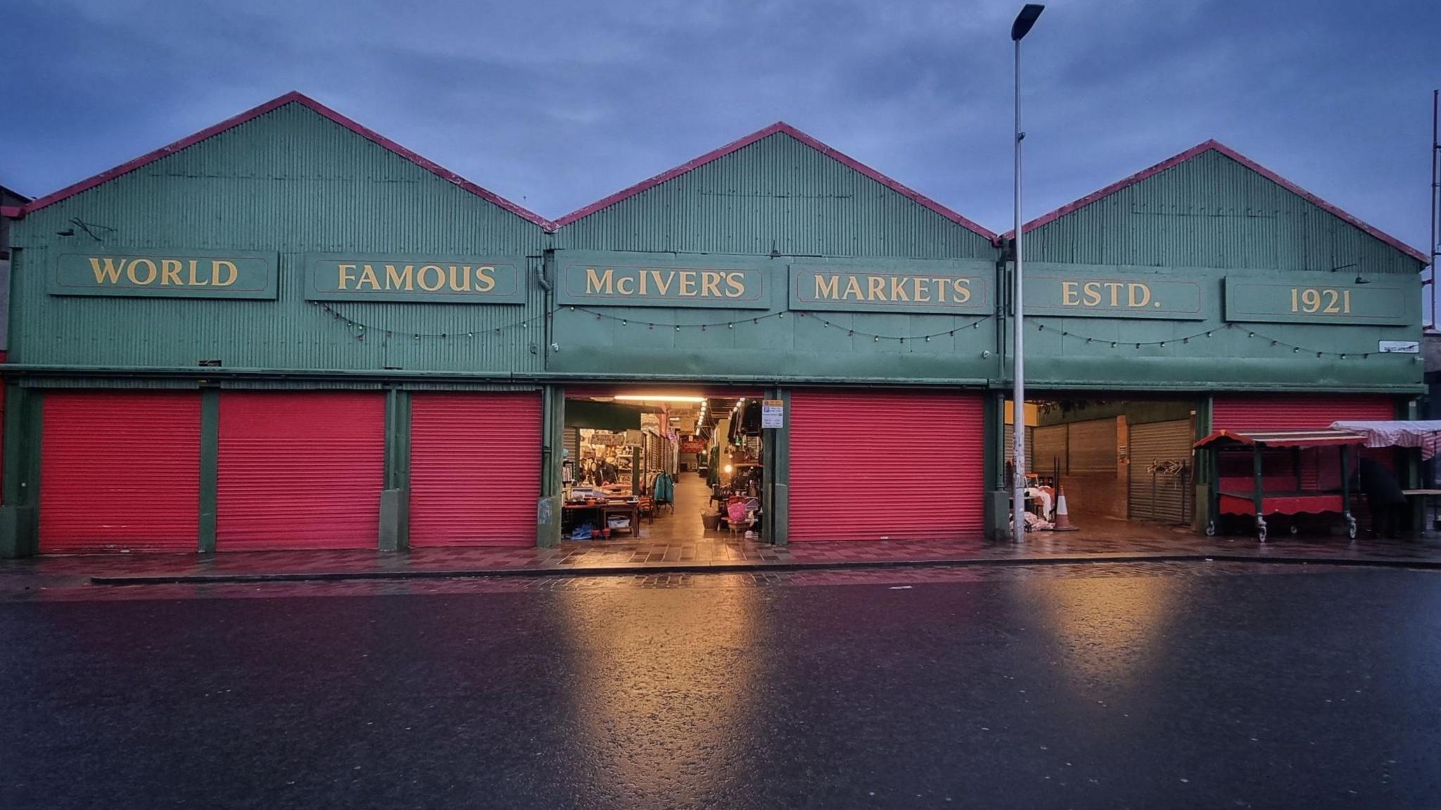 A row of three large sheds. Along the top of the sheds about red shuttered doors are signs reading: "World famous McIver's markets. Estd. 1921". Two of the doors are open and light spills on to a wet road.