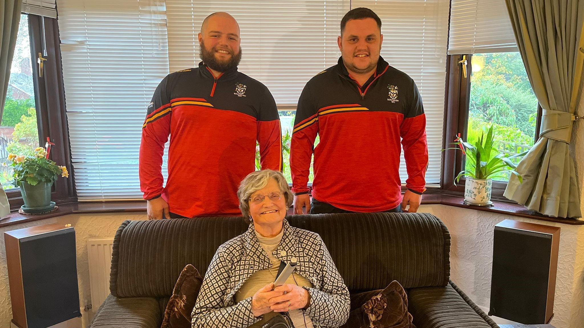 Two men in rugby uniform stand behind a sofa in a living room, and an elderly woman smiles at the camera, sitting on the sofa holding a TV remote 