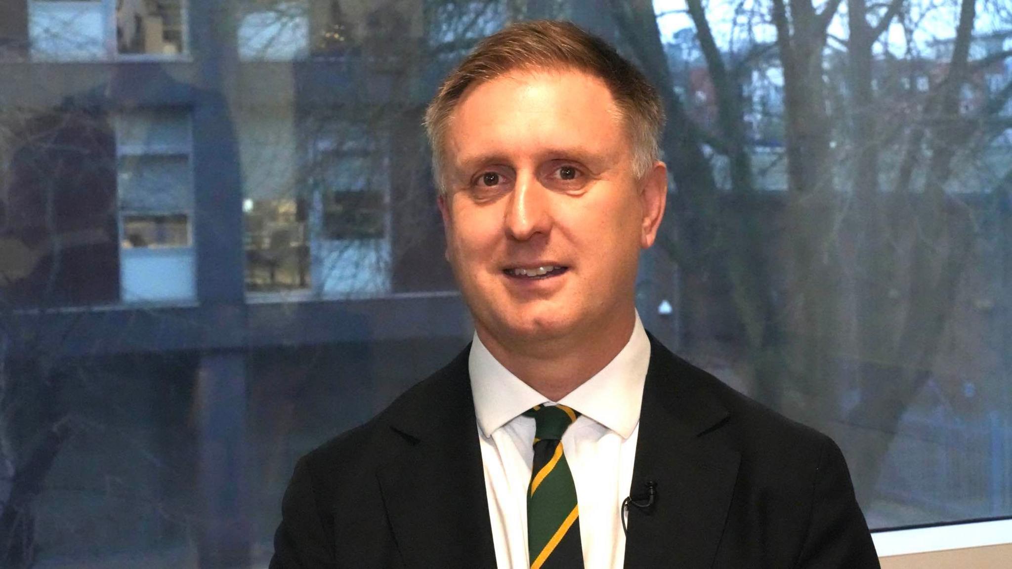 Ed Garratt smiles at the camera in front of a large window. He wears a black suit with a green and yellow tie. He has short grey hair.