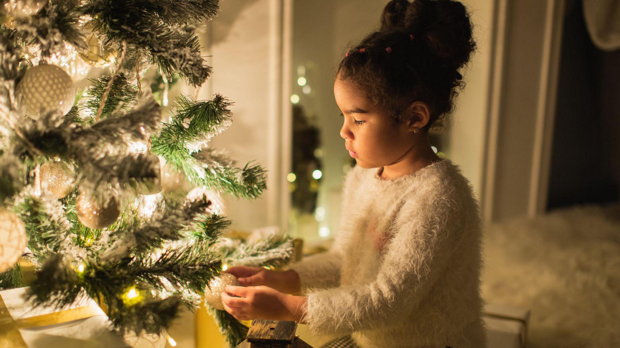 A girl with brown curly hair tied up in a bun wears a white fluffy jumper and hangs a decoration on a Christmas tree with white glowing lights on it