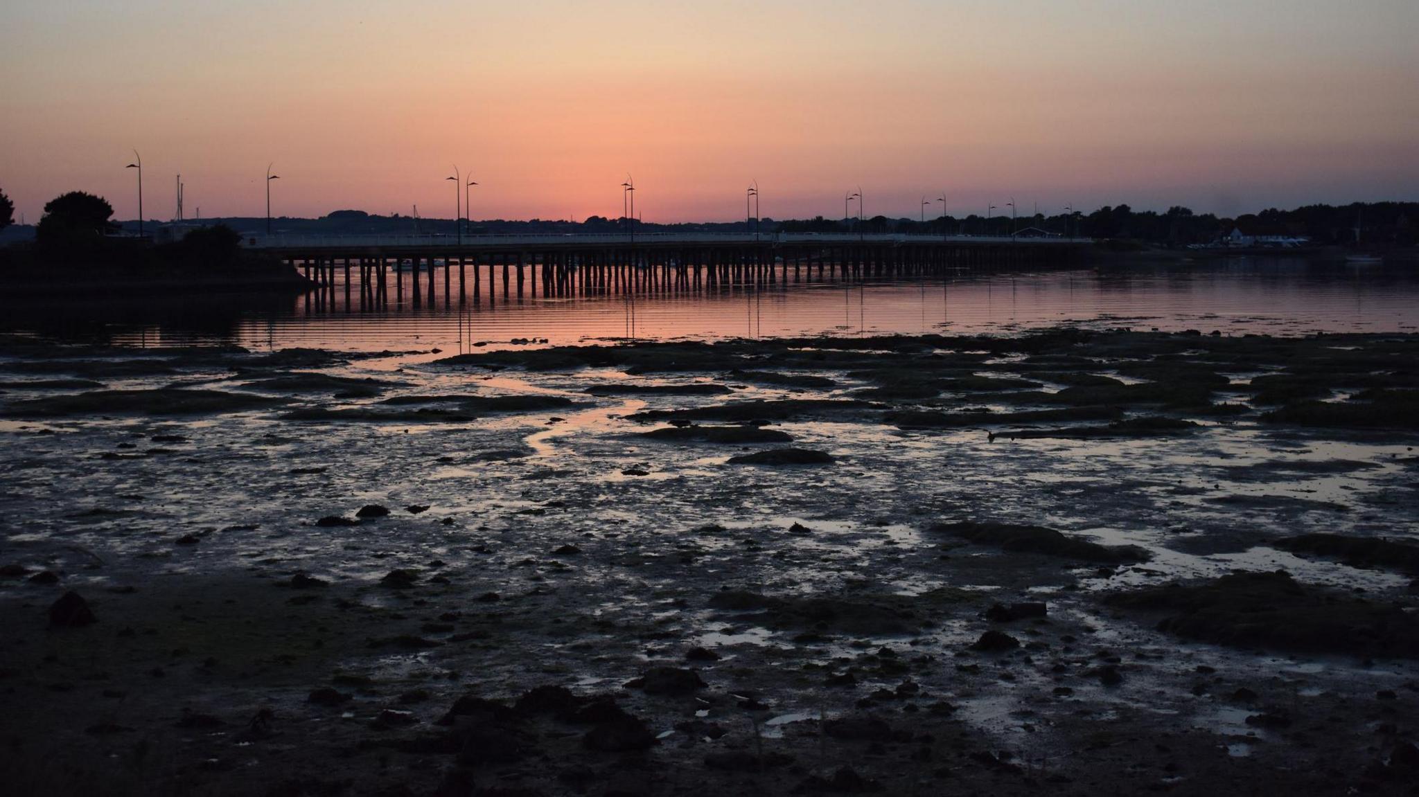 Langstone Harbour at sunset. Mud on the shoreline is illuminated by a red sunset. The land in the background is in silhouette