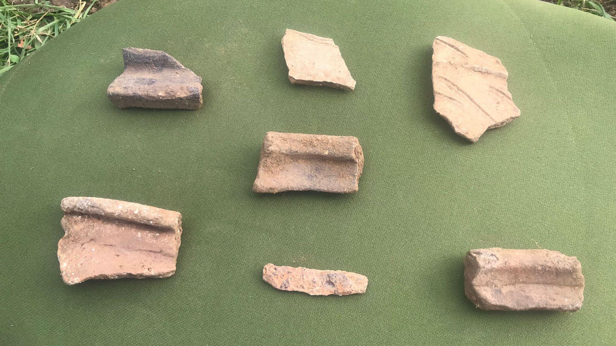 Seven fragments of medieval pottery spaced out on a green piece of fabric, with soil and grass seen underneath it