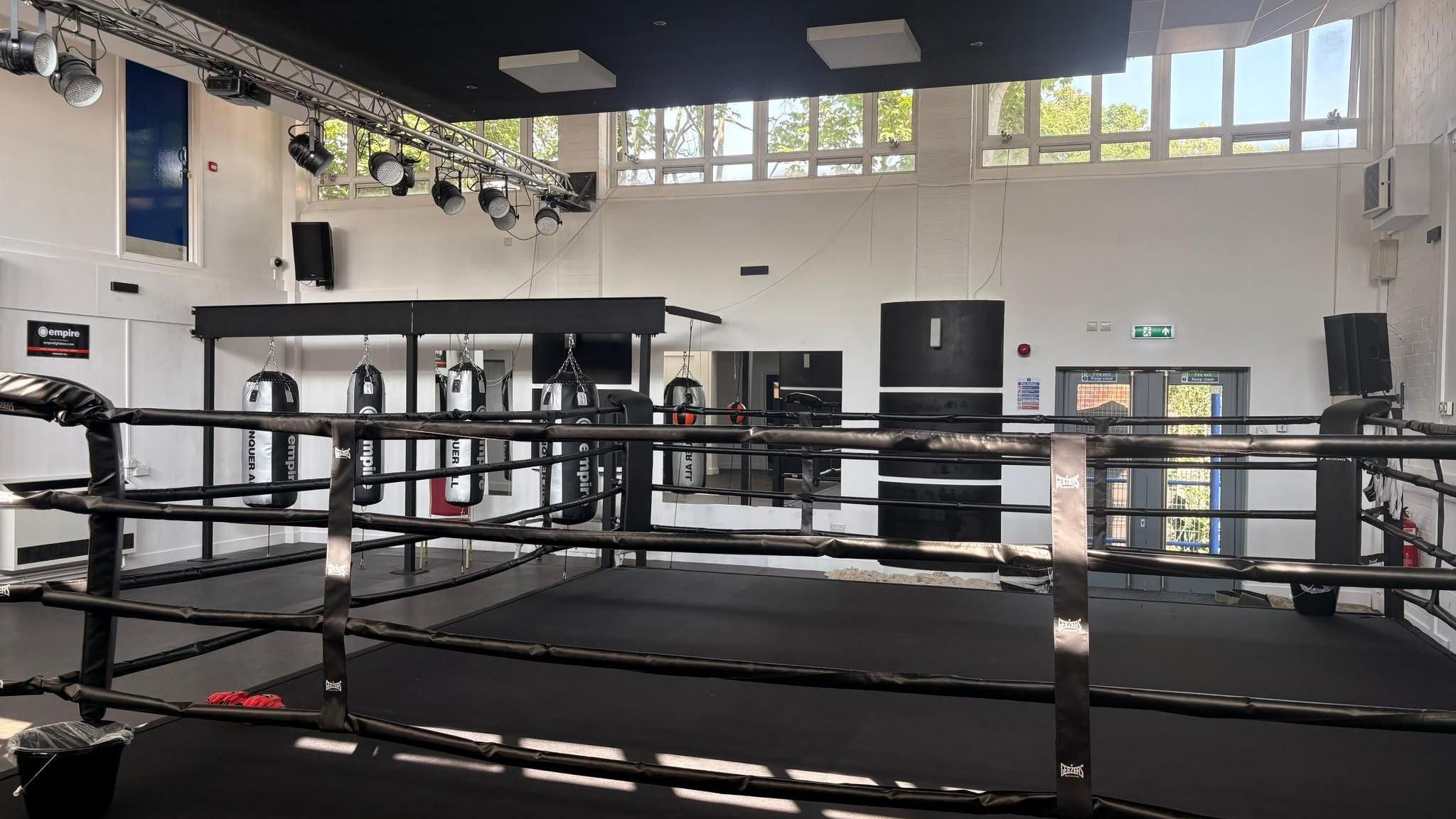 A spacious boxing gym with a large black boxing ring in the foreground. Several heavy punching bags hang in the background beneath a metal lighting rig fitted with stage lights.