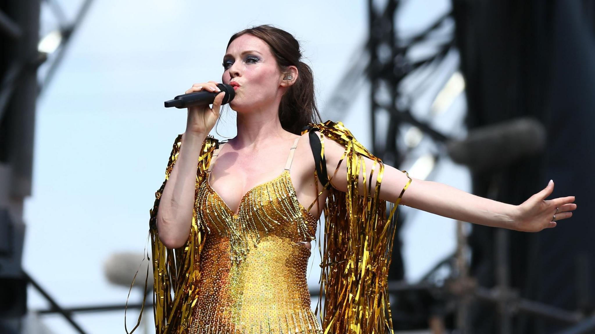 Sophie Ellis-Bextor wearing a gold outfit on stage, covered in gold streamers. she is singing into a microphone and has an outstretched arm.