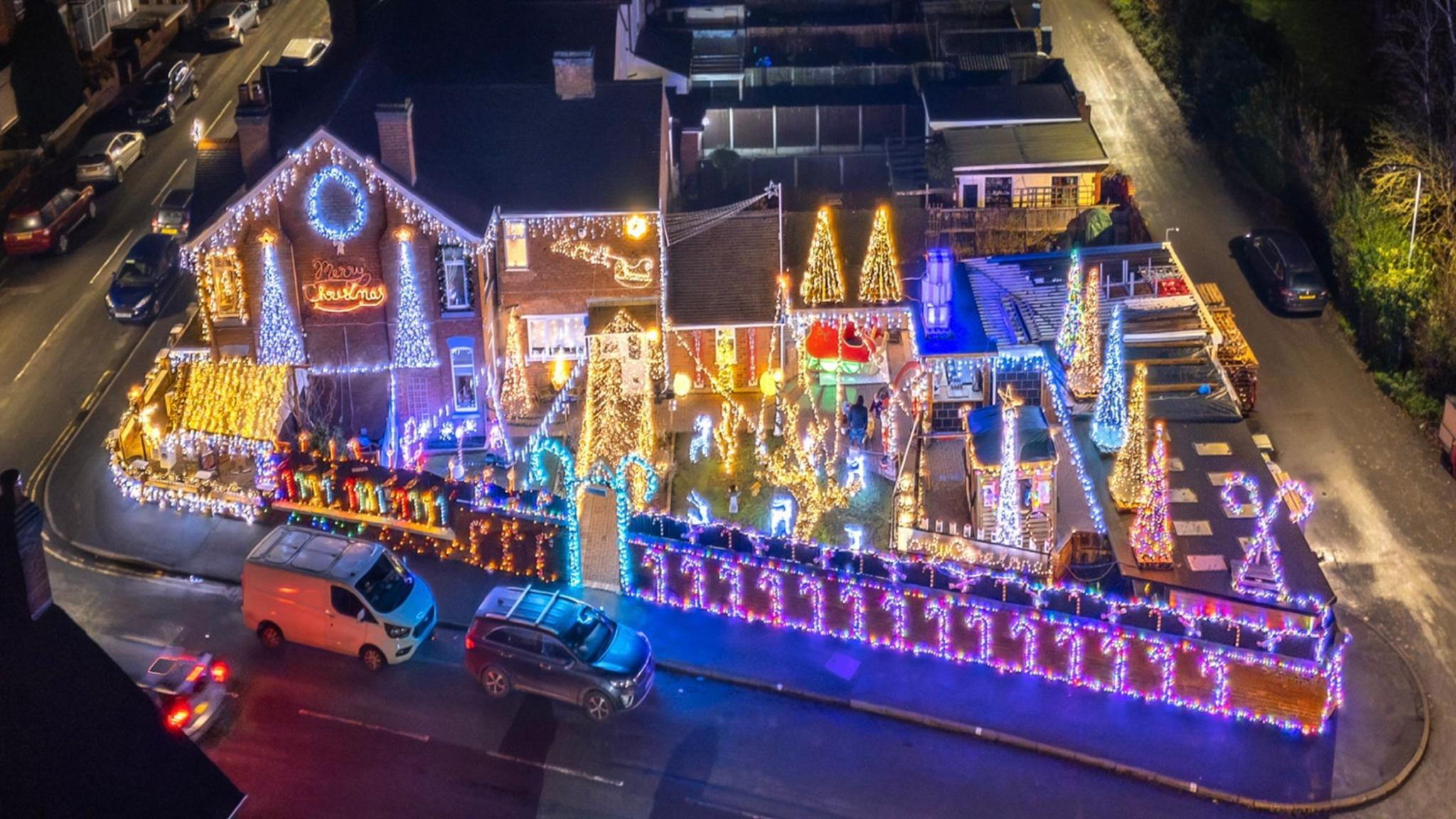 An aerial shot of an end terrace house and garden at night. The house and garden are heavily decorated by Christmas themed lights. There are illuminated trees, candy canes, stars and reindeer. There are a few cars and vans on the street.