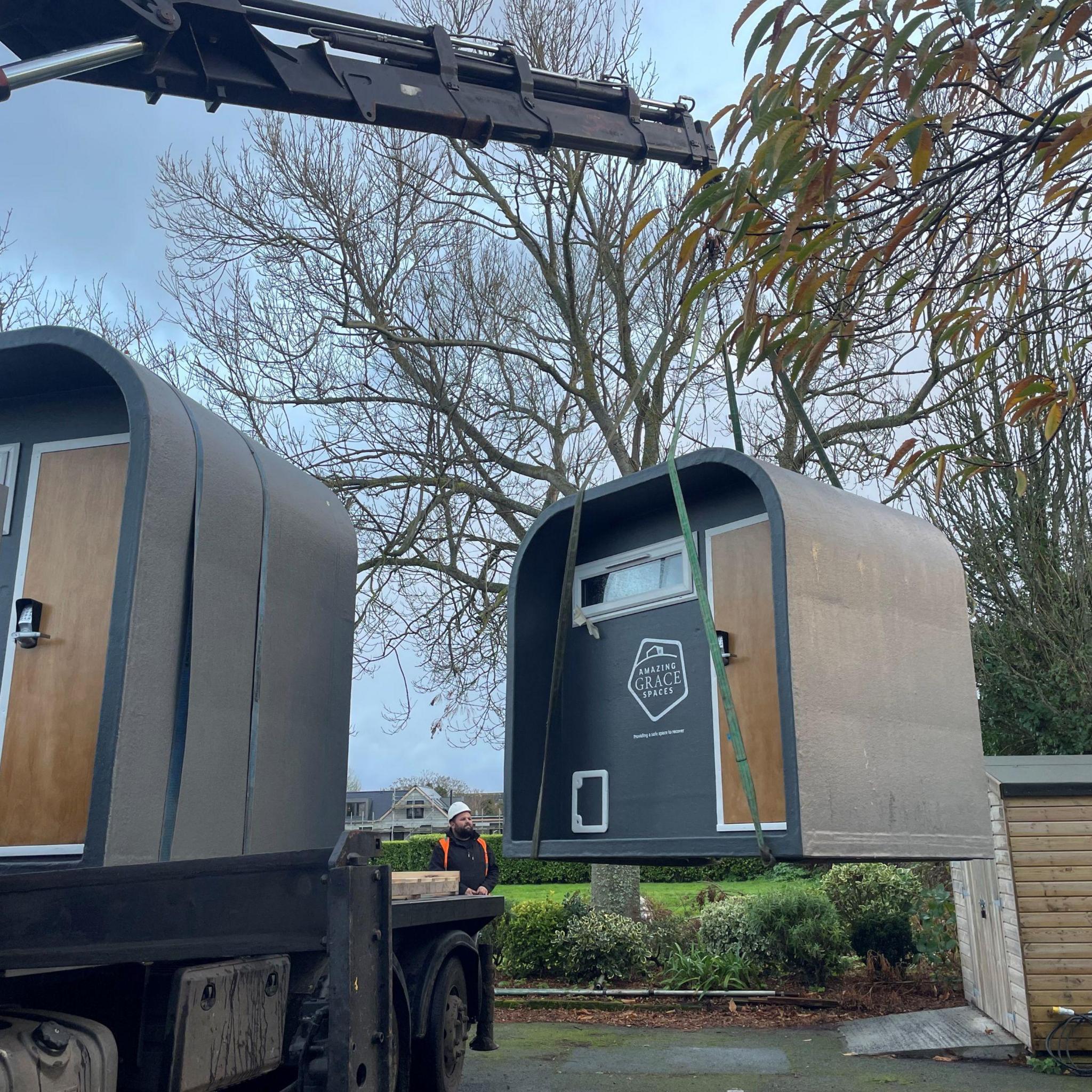 A grey pod a small white window and brown door with keypad is being moved from a lorry to the ground by a crane arm on the lorry. It has an Amazing Grace Spaces logo on the front.