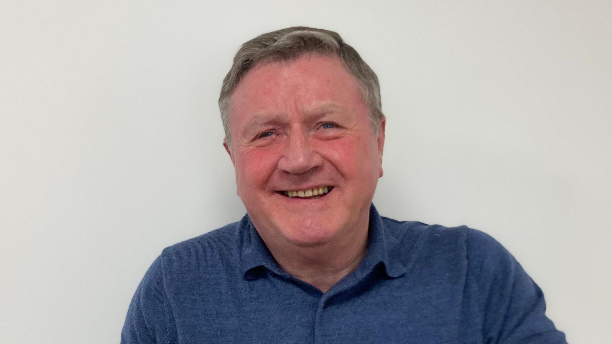 Dennis Jones smiling at the camera against a white wall, wearing a blue polo shirt.