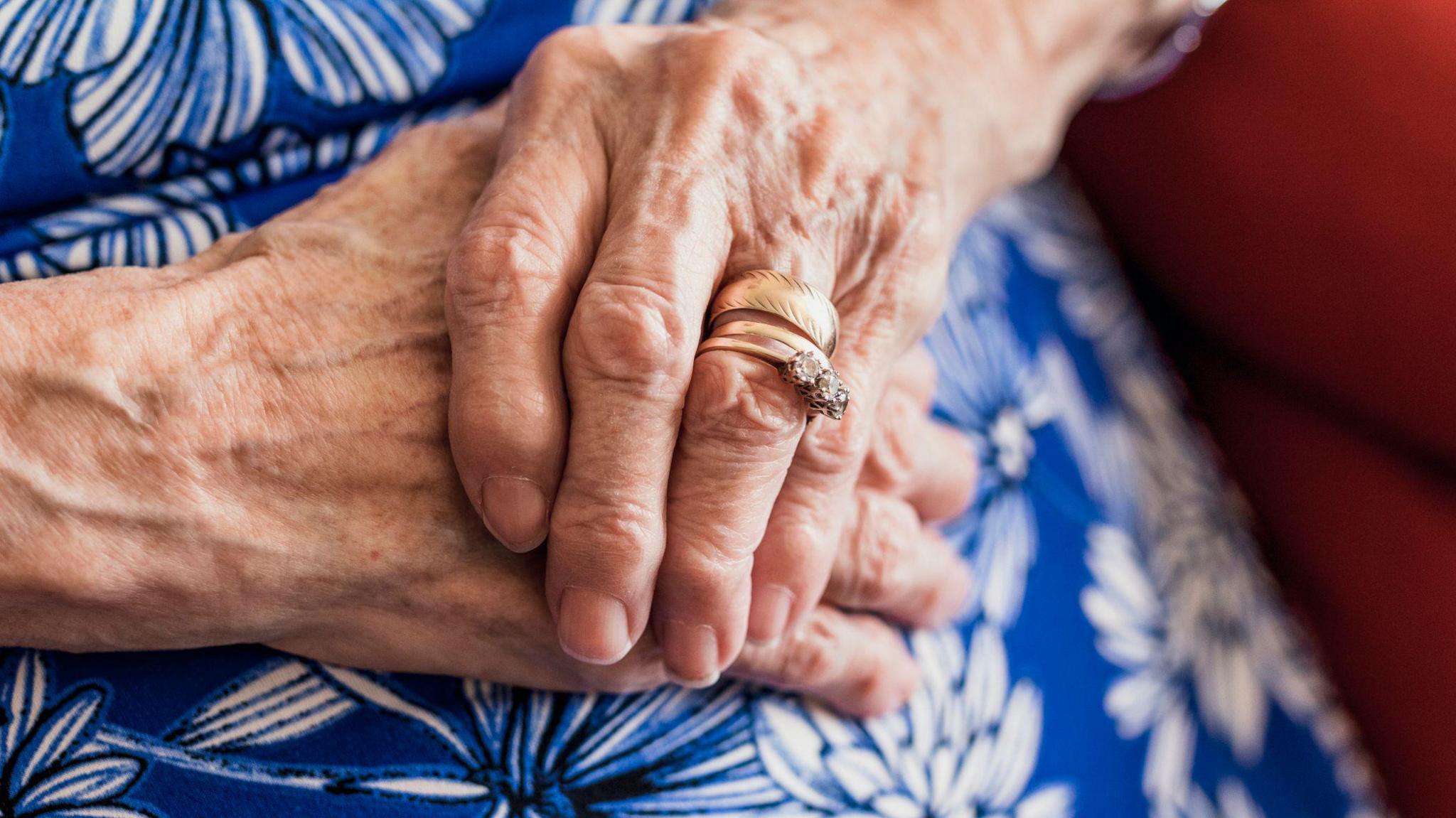 An elderly woman's hands resting on her lap. She wears a blue floral dress and a wedding ring.